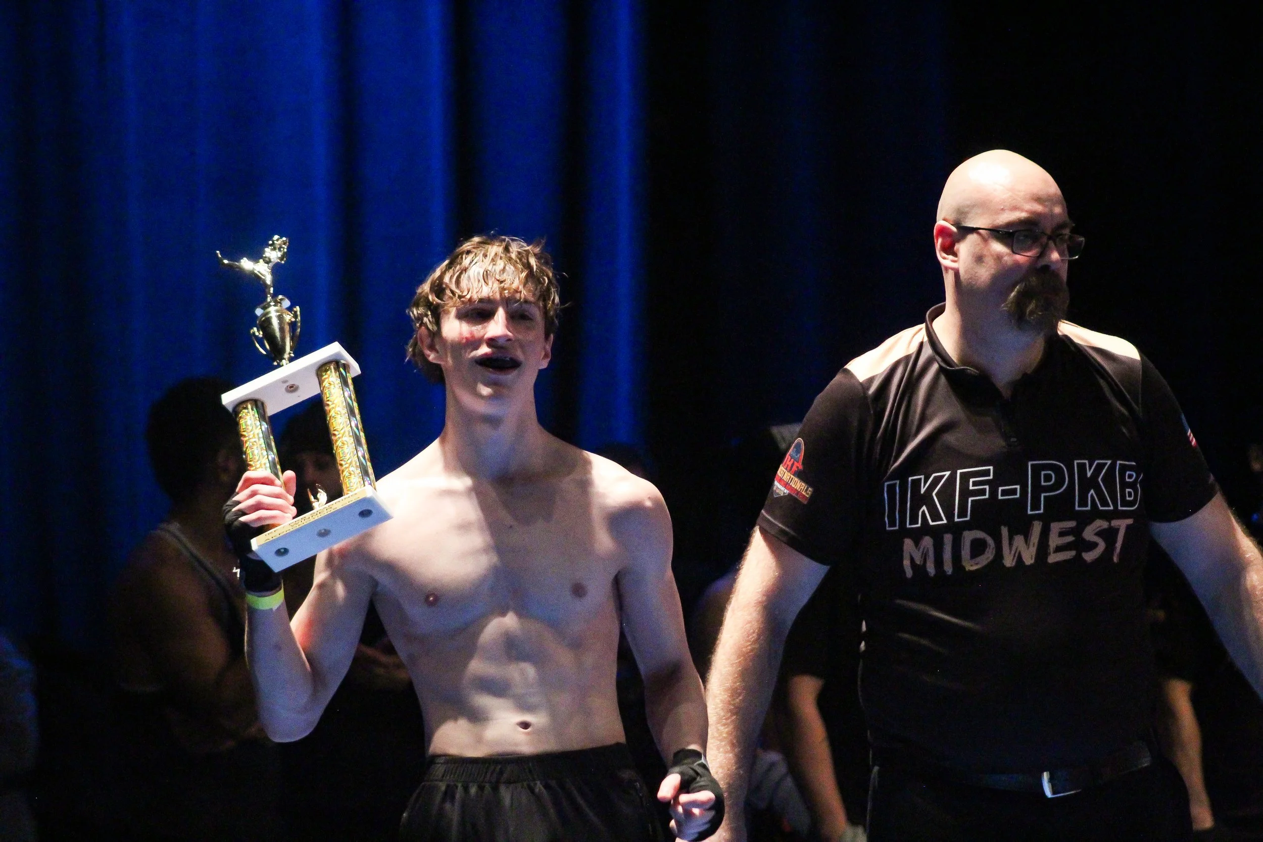 A young male athlete with curly hair and no shirt holding a trophy, standing next to a man in a black shirt with 'IKF-PKB MIDWEST' written on it, amid a dark background with blue curtains.
