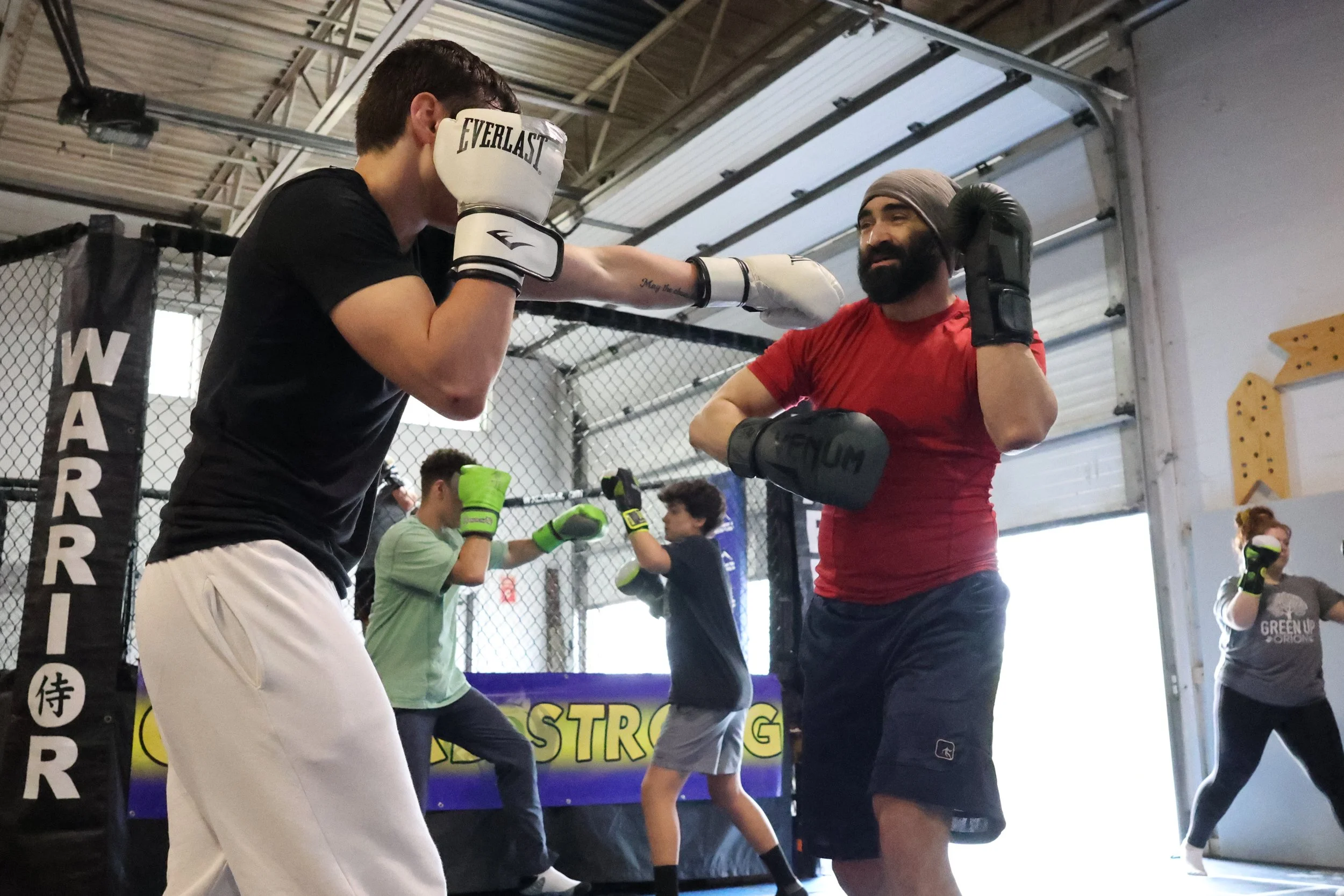 Two men sparring in a boxing gym, with other people training in the background, showcasing a boxing or martial arts training session.