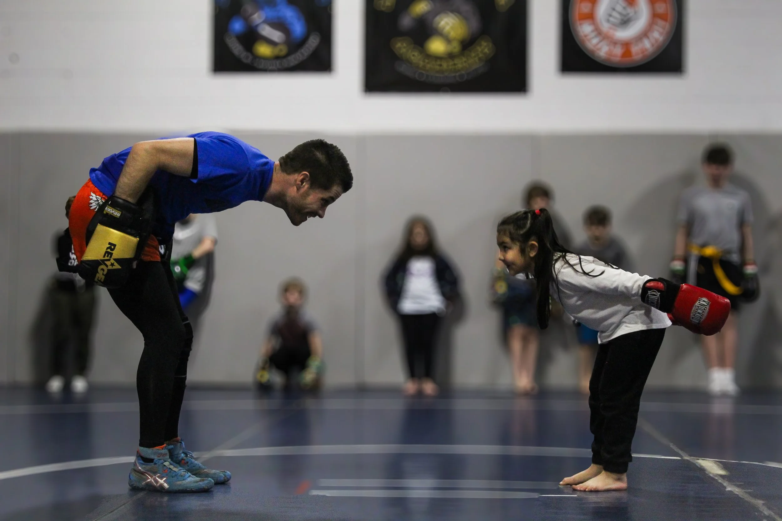 An adult male and young girl practicing martial arts, bowing to each other in a dojo with children in the background.