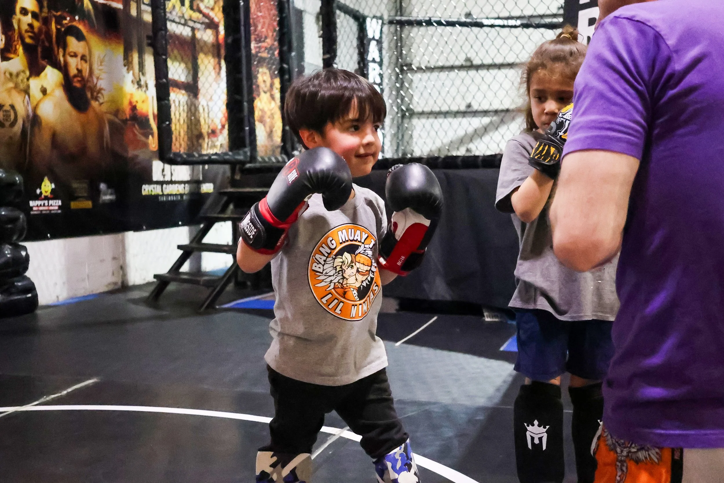 A young boy wearing boxing gloves, a gray t-shirt with 'Bang Muay Lil Ninjas' logo, and camouflage knee pads practicing boxing with an adult trainer in a gym with a UFC poster in the background.