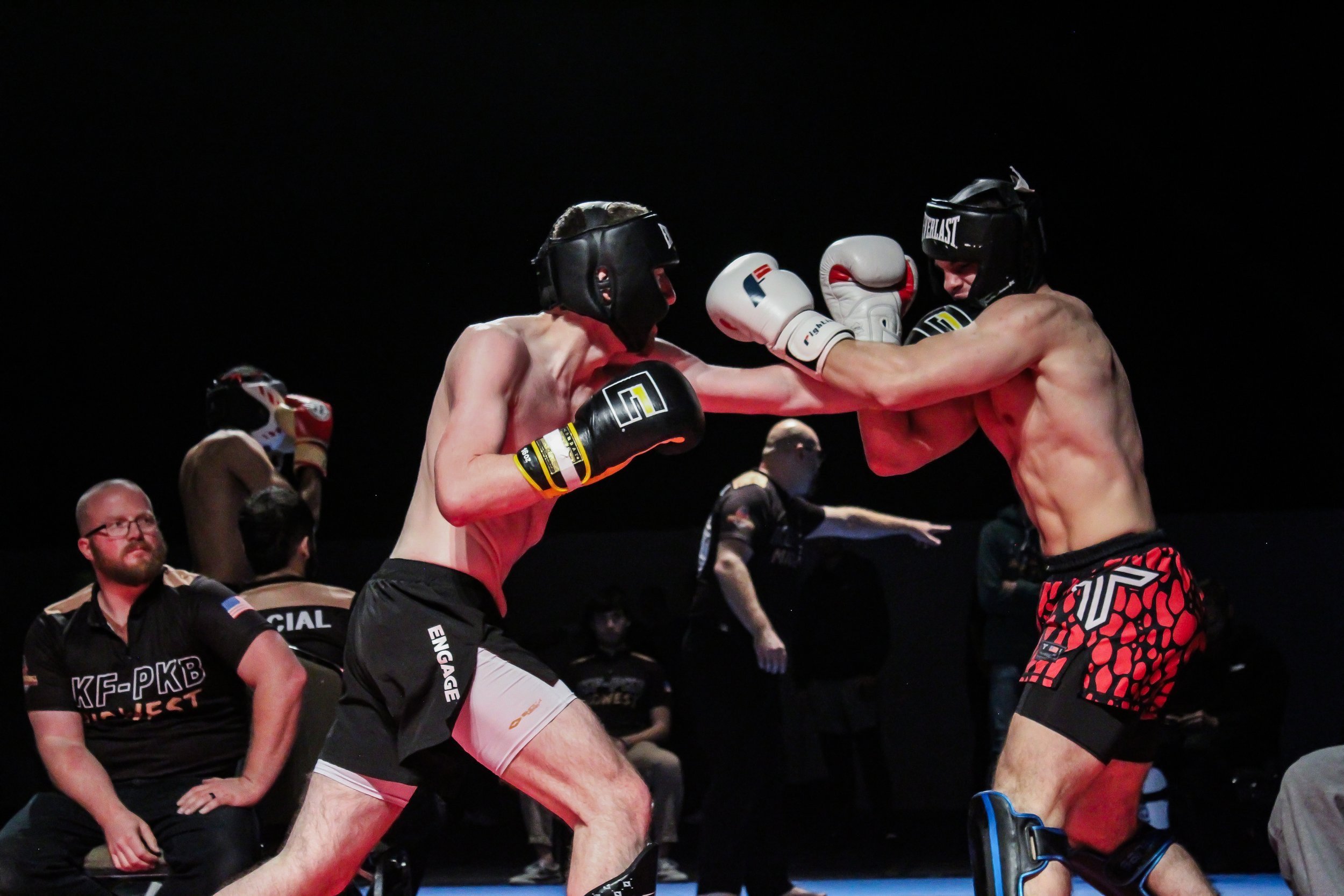 Two shirtless male fighters wearing headgear and gloves engaged in a boxing or kickboxing match, with one delivering a punch to the other's face. Several officials and audience members observe in the background.