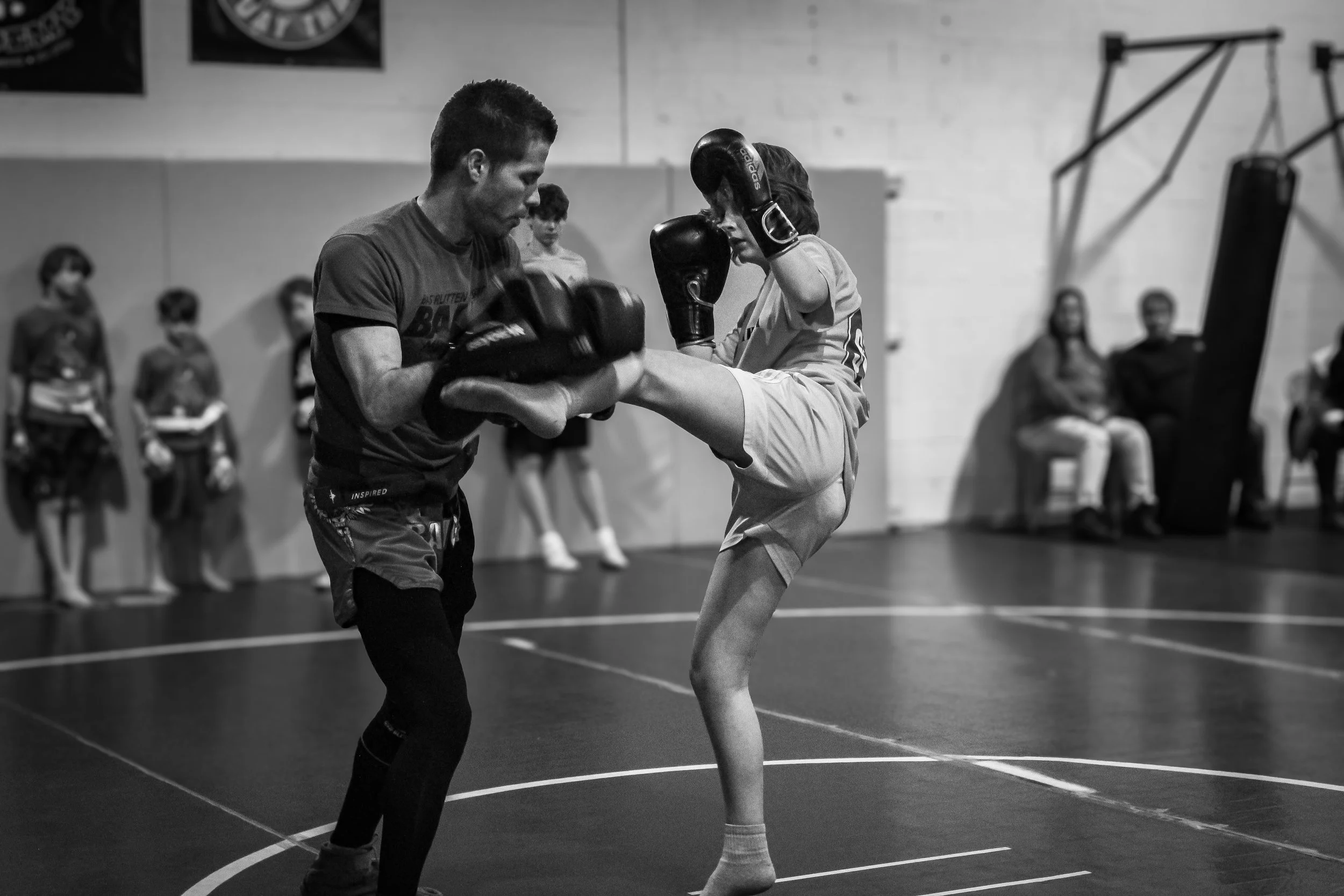 A martial arts training session in a gym where a young girl is practicing a high kick with a trainer holding pads, and several people sitting and watching in the background.