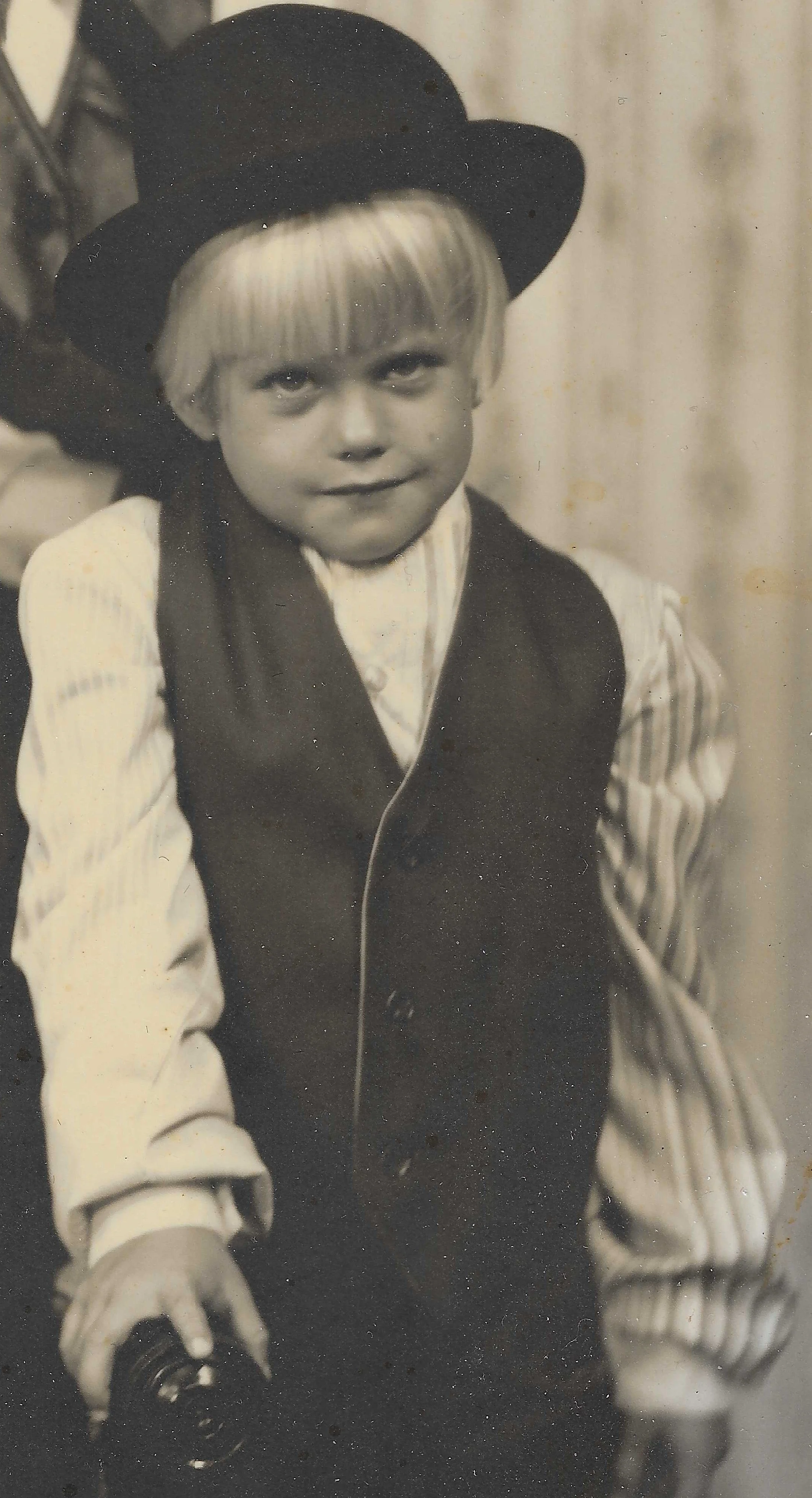 A young boy dressed in vintage clothing, wearing a striped shirt, vest, and a bowler hat, looks at the camera with a slight smile.