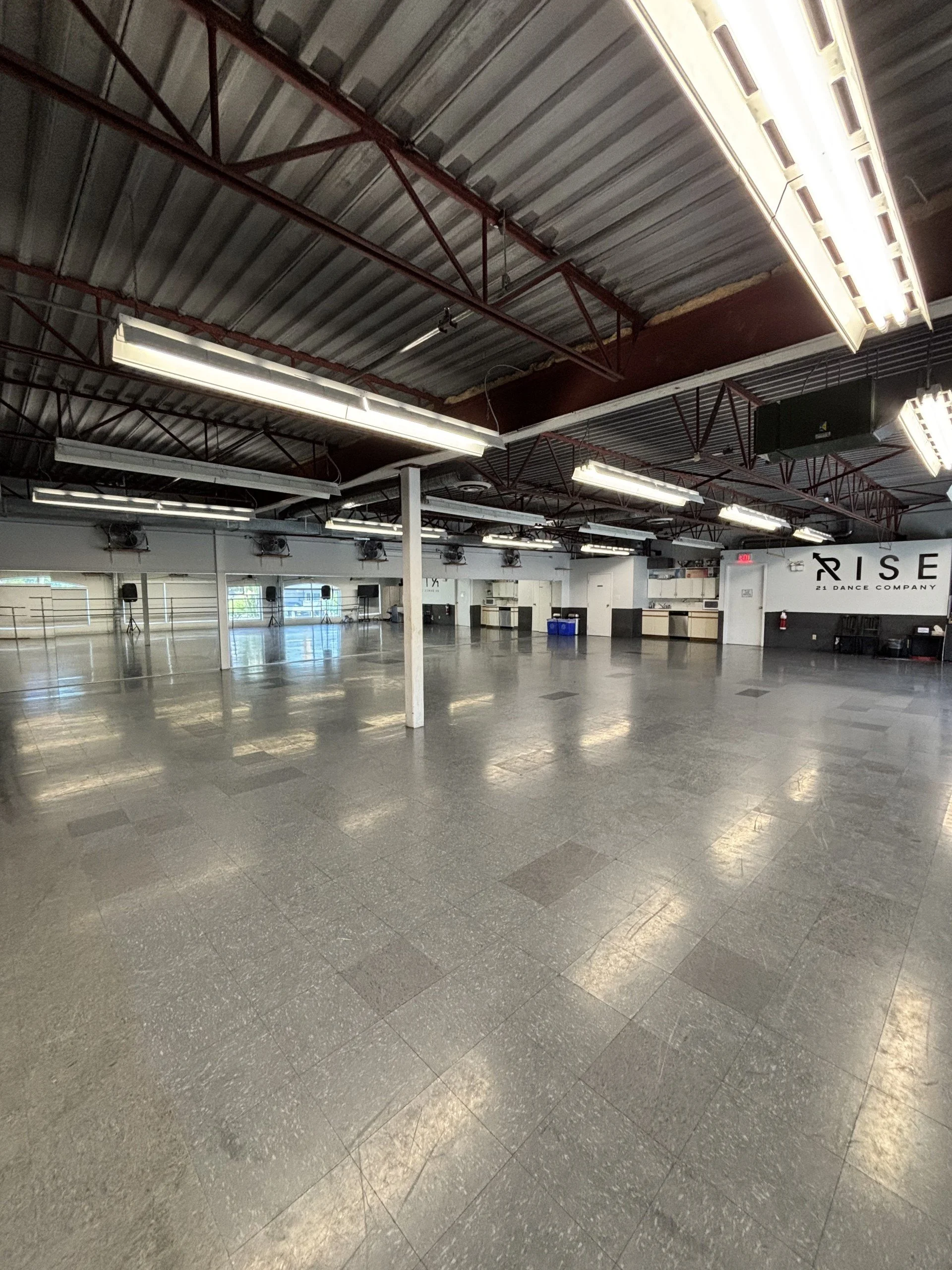 Empty dance studio with large mirrors, ballet barres, and fluorescent lighting fixtures on a metal ceiling. The floor is polished and reflective, with some equipment and a wall sign that reads 'RISE 21 Dance Company' visible in the background.