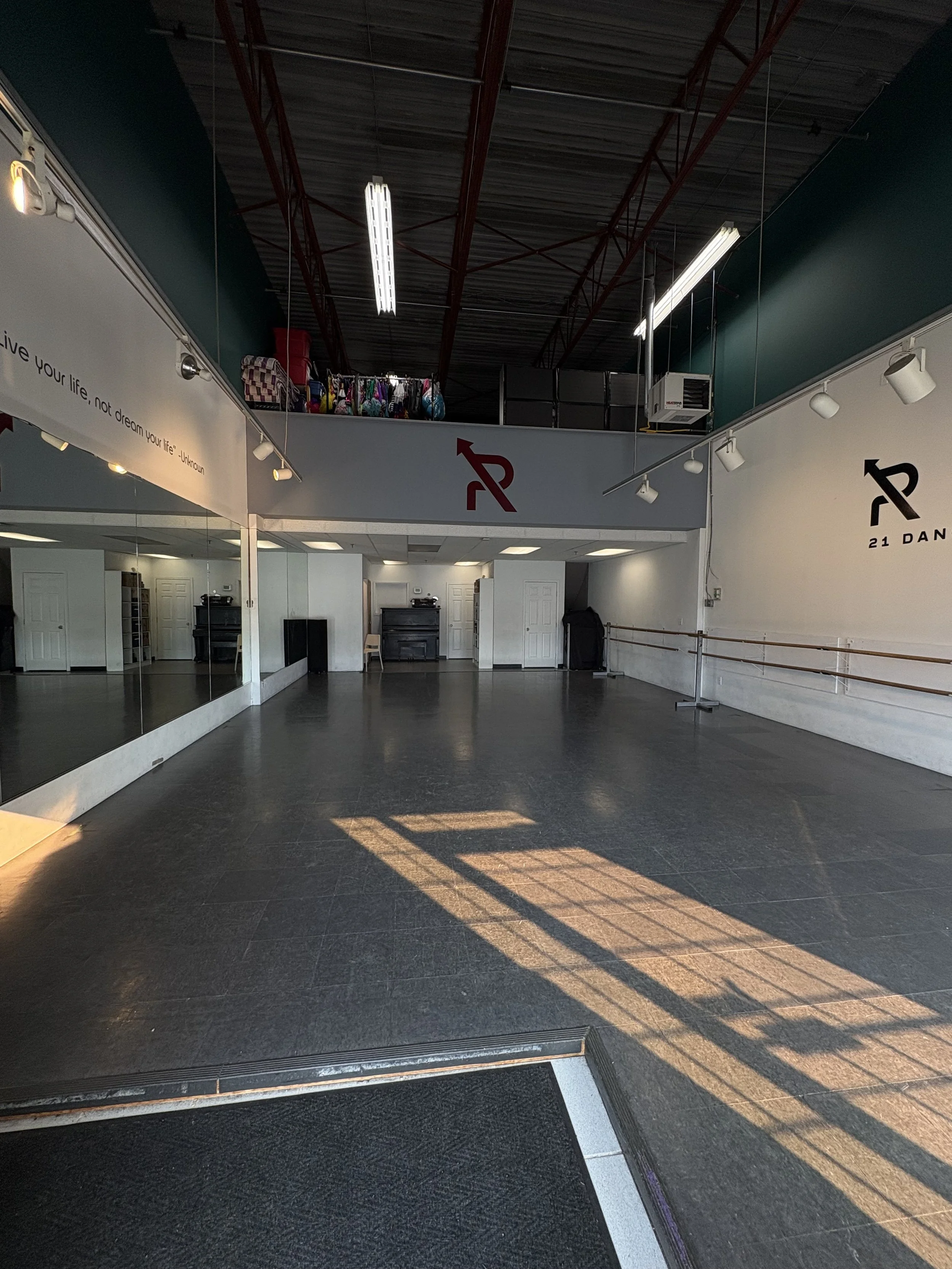 An empty dance studio with a wooden floor, mirrors lining the left wall, a ballet barre on the right wall, and a loft area with bags and belongings. The studio is lit with ceiling lights and sunlight casting shadows on the floor.