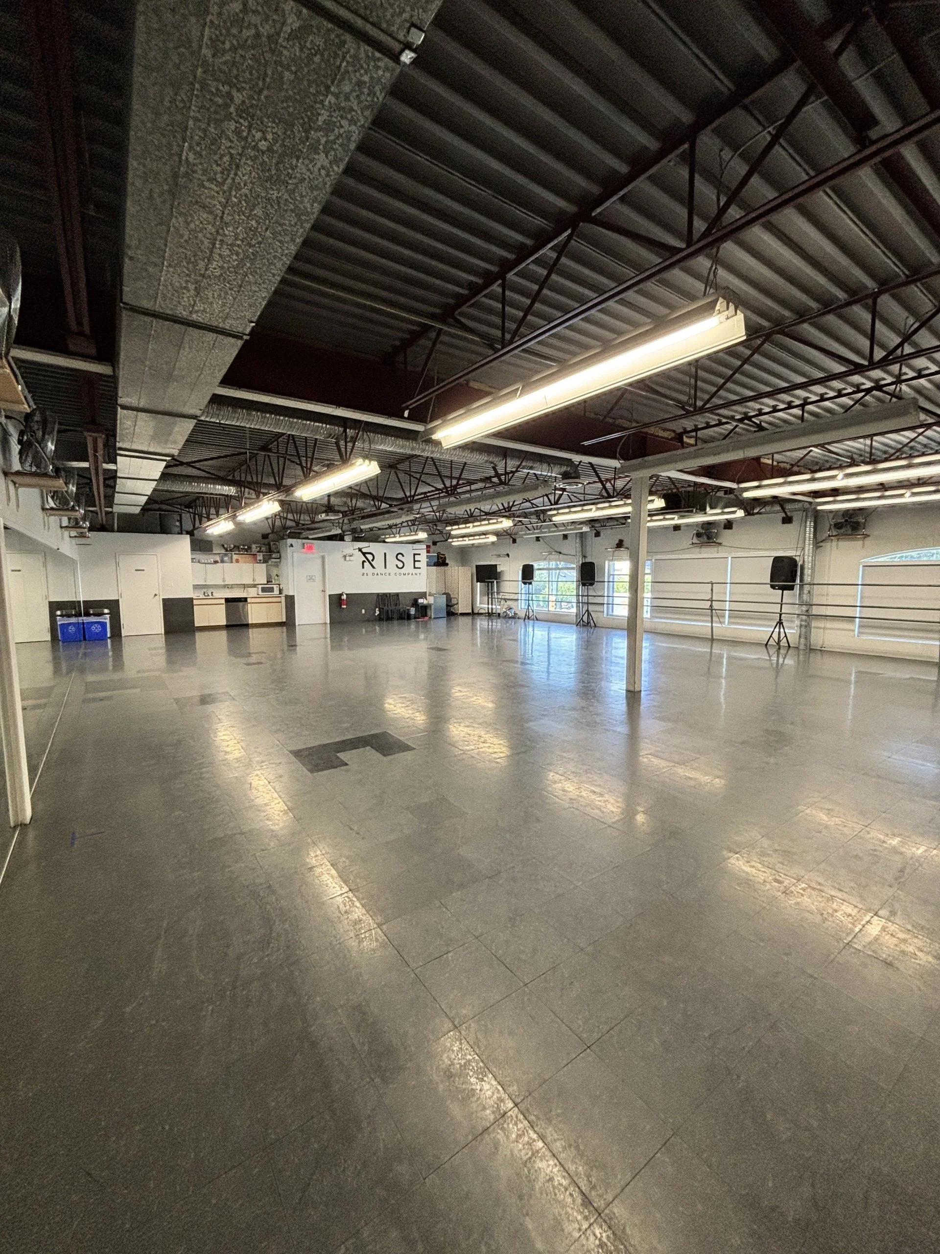 Empty dance studio with large mirrors on the left wall, ballet barres on the right, and a sign that says 'RISE 21 DANCE COMPANY' in the background. The room has polished floors, fluorescent lighting, and windows on one side.