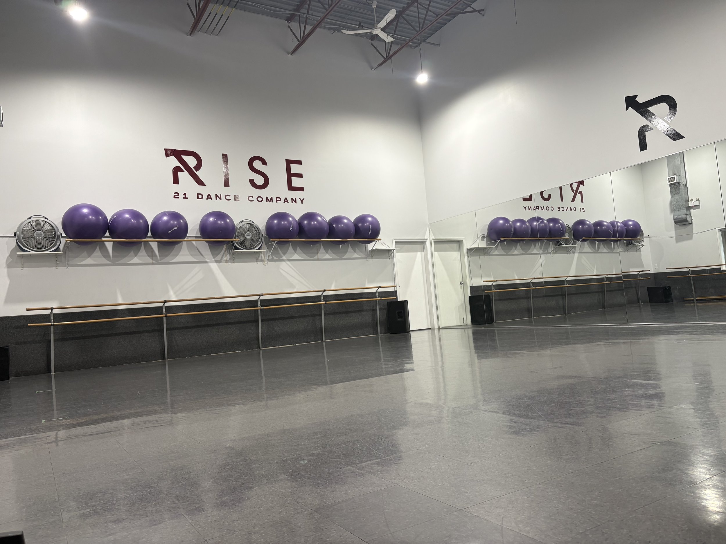 Empty dance studio with a large mirror on the right wall, ballet barres, purple exercise balls on a shelf, and a sign that reads 'RISE 21 DANCE COMPANY' on the back wall. The studio has a gray floor and a high ceiling with exposed structural elements and ceiling fans.