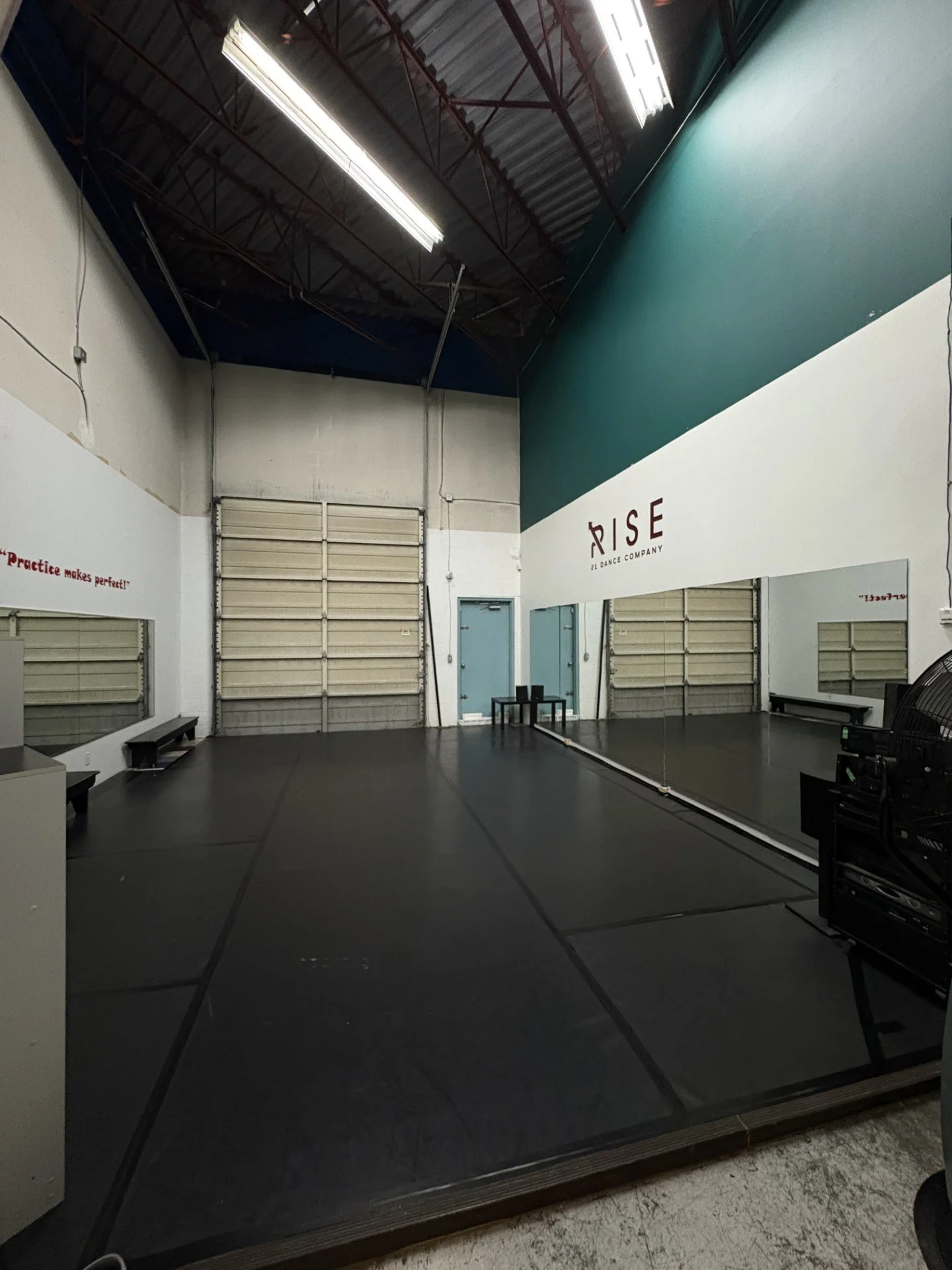 A dance studio with black flooring, mirrors on one wall, and a large roller door at the back. The studio has high ceilings with exposed metal beams and bright overhead fluorescent lights. There are benches and a fan on one side, and the studio walls have motivational quotes and branding.