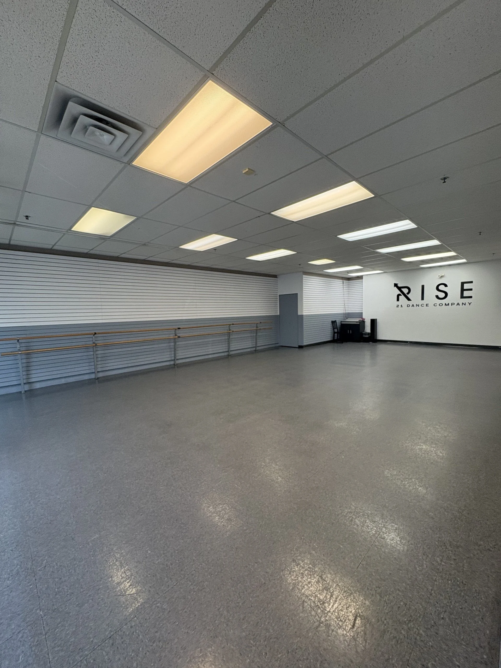 Empty dance studio with gray flooring, ballet barres along the wall, fluorescent ceiling lights, and a sign reading 'RISE 21 DANCE COMPANY' on the back wall.