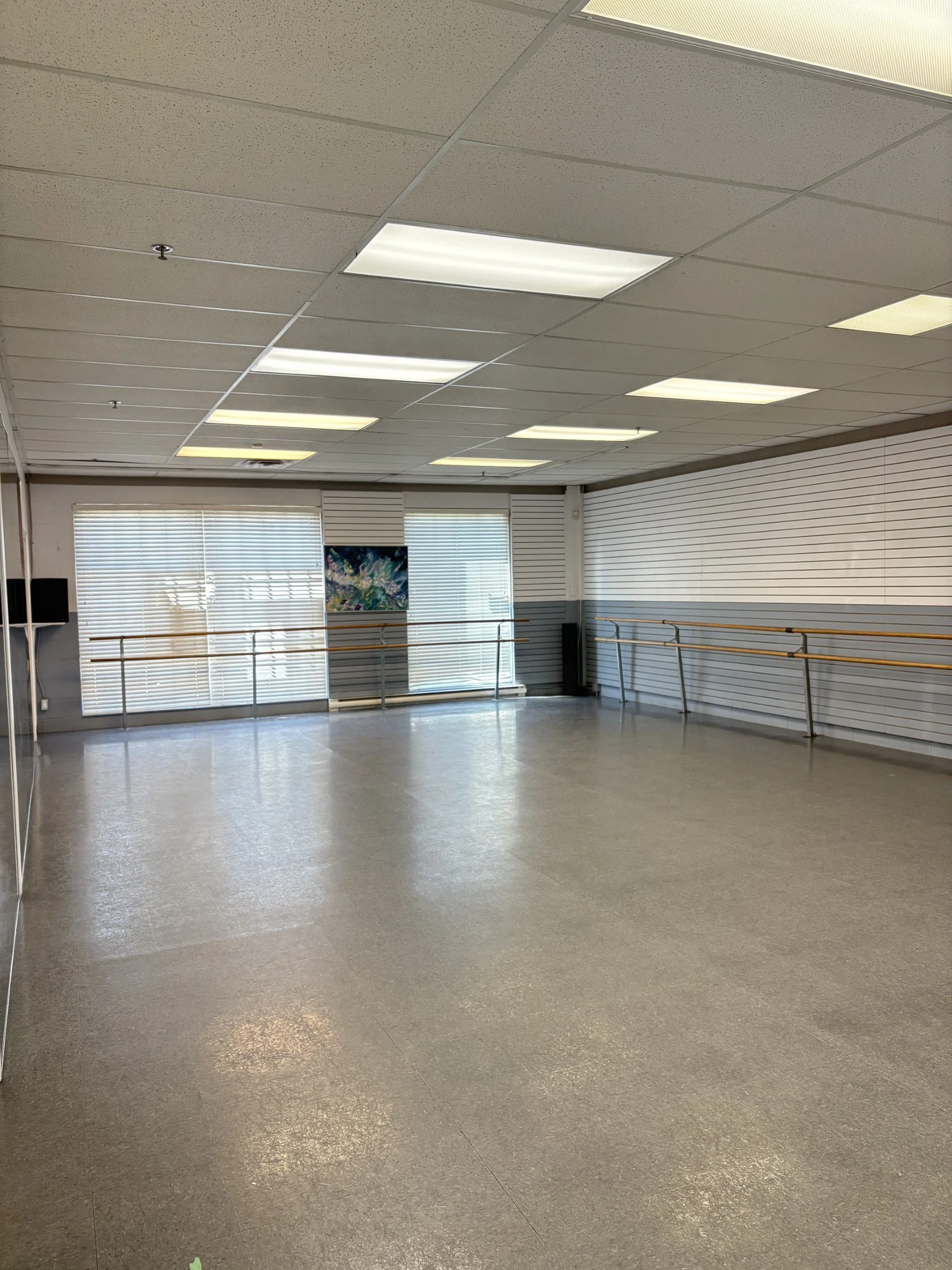 Empty dance studio with mirrored wall, ballet barres, and large windows with blinds.