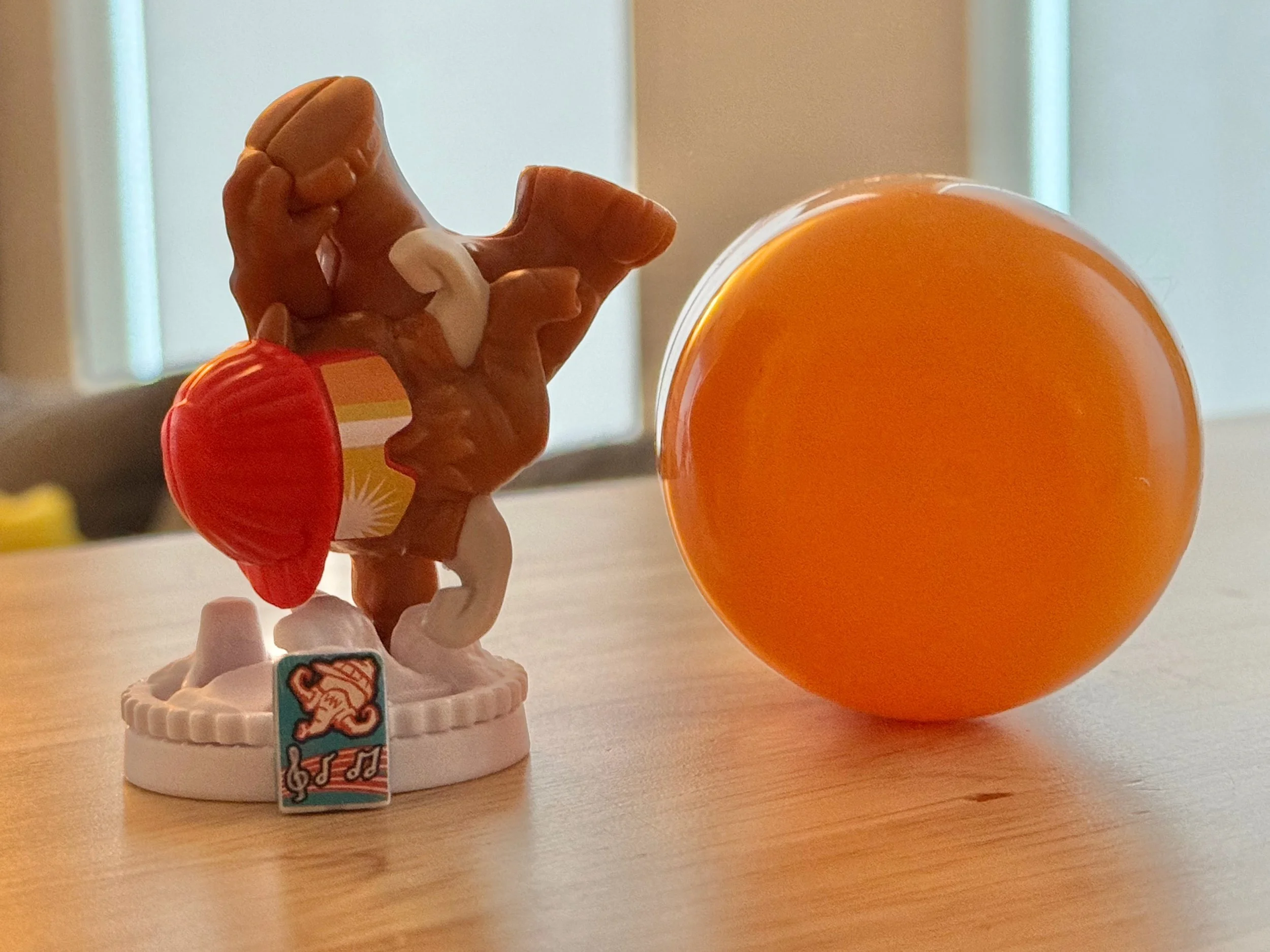 A Kinder Surprise woolly mammoth figurine posed in a breakdancing move on a snow base, wearing an orange toque and orange sunglasses with yellow lenses, with the orange Kinder Surprise egg placed to the right.