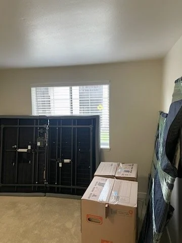 Empty room with a window, two cardboard boxes, and a disassembled black TV stand or furniture piece leaning against the wall.