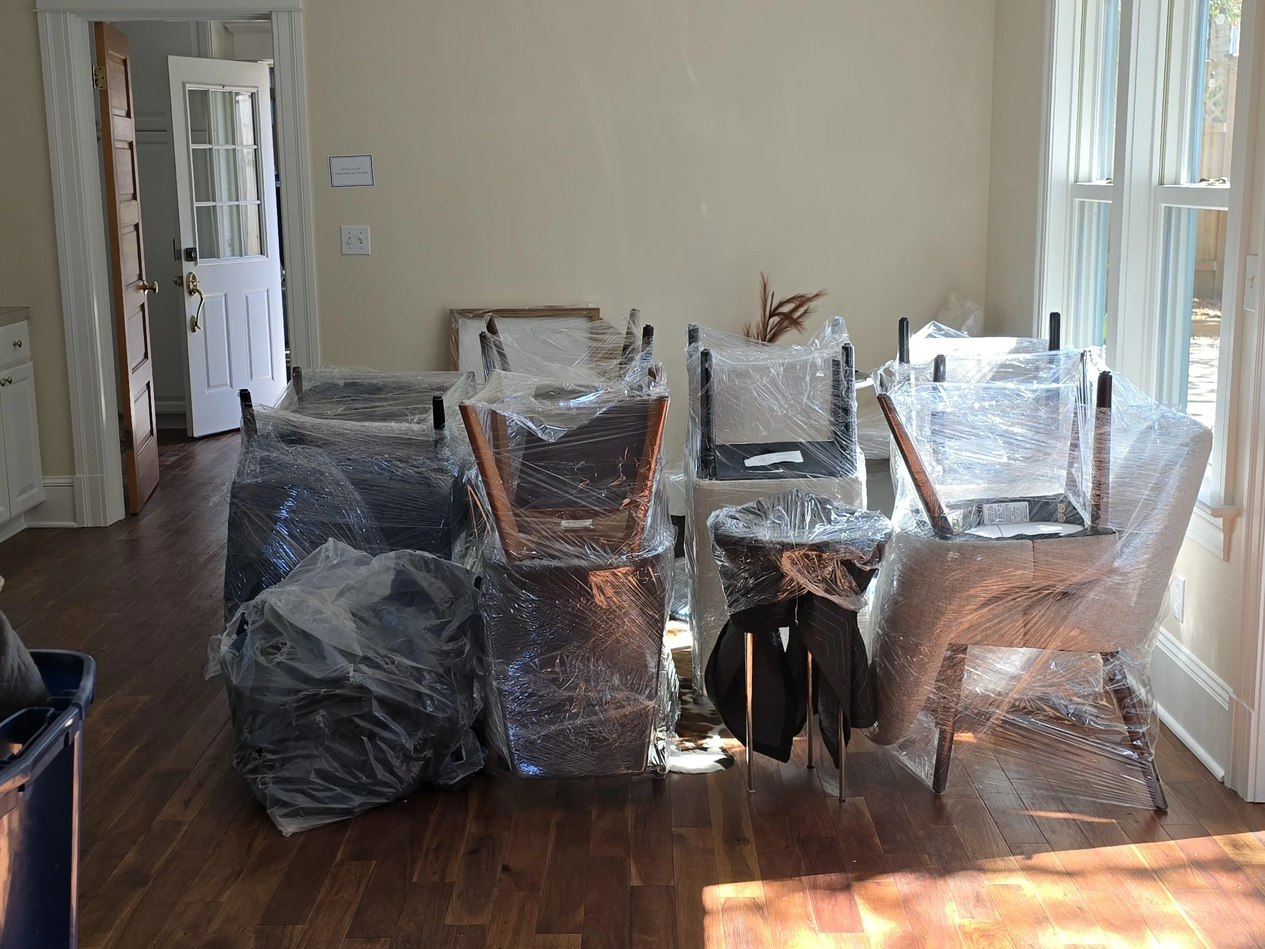 Dining room with six chairs wrapped in plastic, some chairs stacked, and a pile of plastic bags on the floor, near a large window and a closed door.