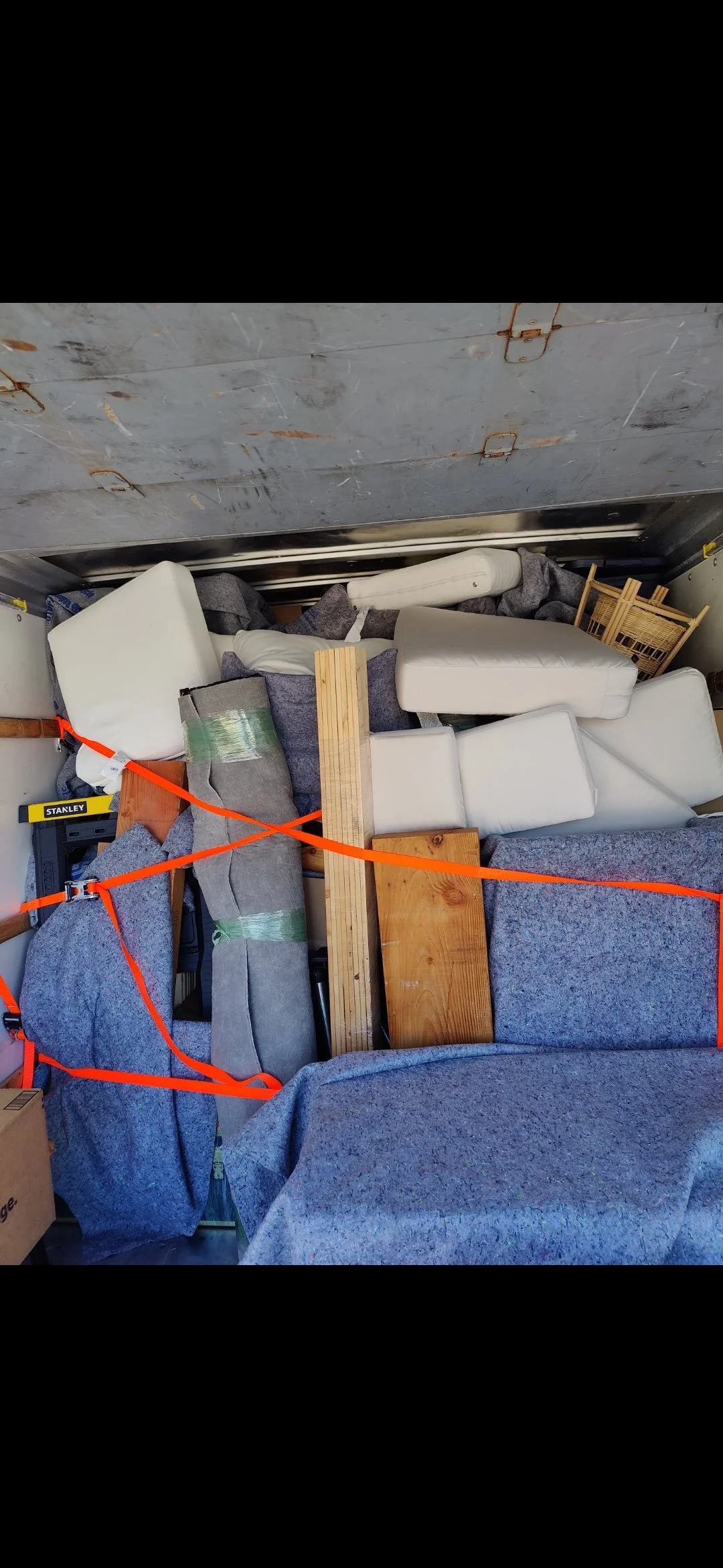 Storage unit filled with various furniture pieces including couches, wooden boards, foam, and fabric coverings, secured with orange straps.