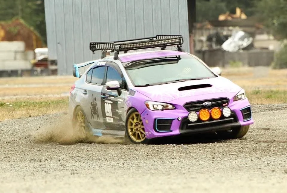 A purple and black rally car with gold wheels driving on a dirt surface, kicking up dust, equipped with additional lights on the front, and a roof rack, in an outdoor setting.