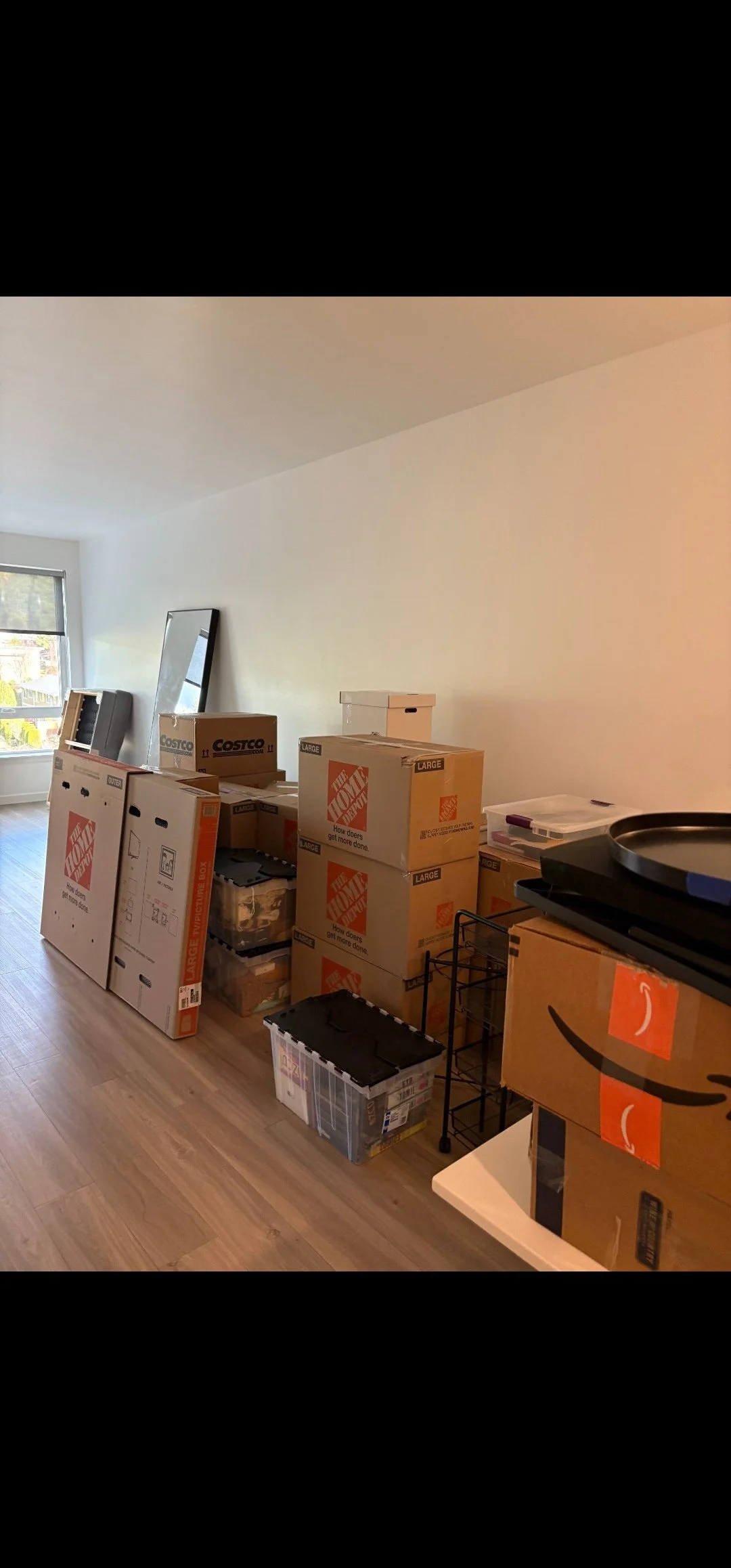 Room with packed moving boxes, including several from Home Depot and Costco, stacked along the wall and on the floor, near a window with blinds, in a room with wooden flooring and minimal furnishings.