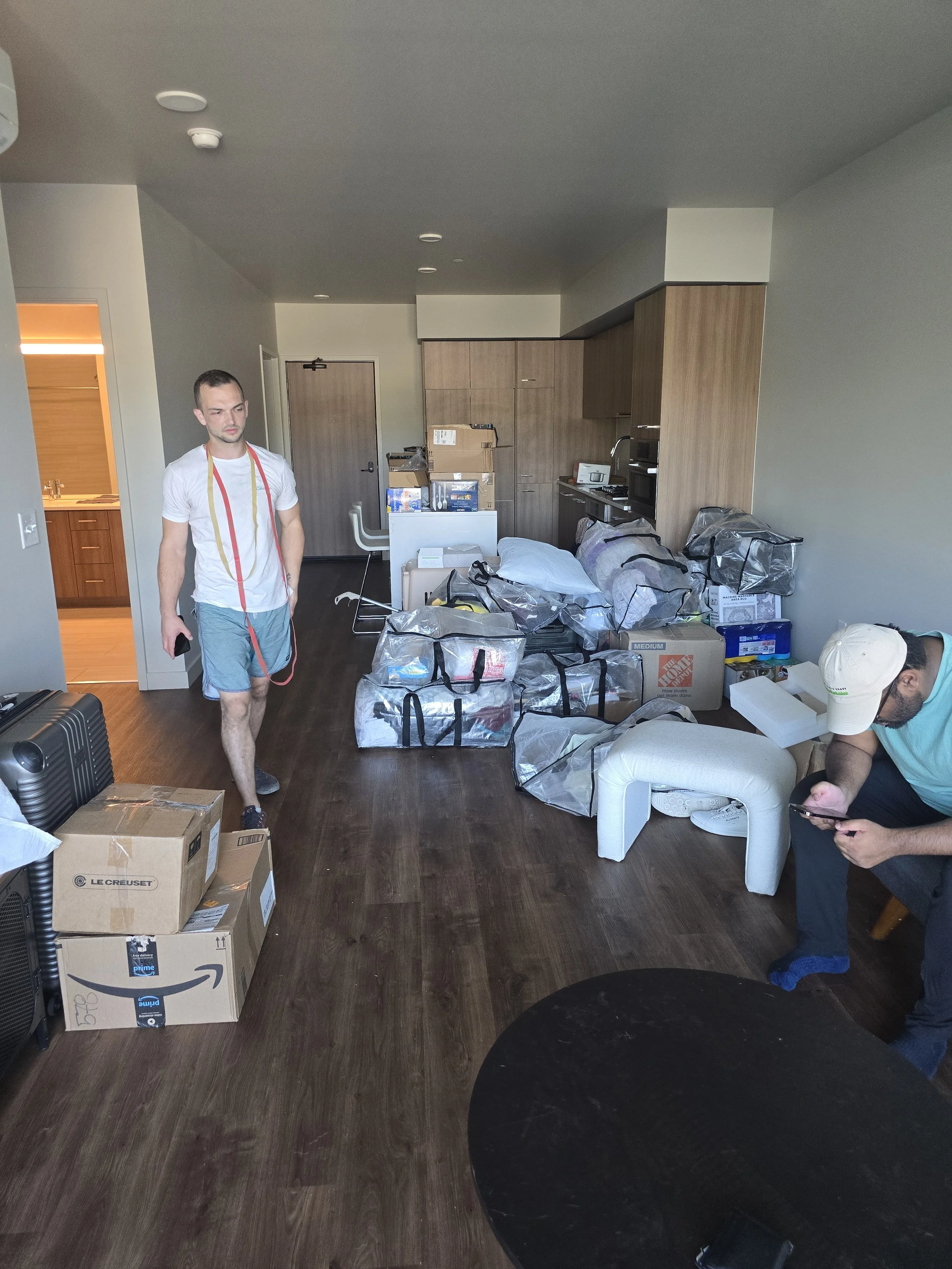 A man in a white t-shirt and shorts walking in a cluttered apartment, with boxes and bags on the floor, and another person sitting on a chair using a phone.