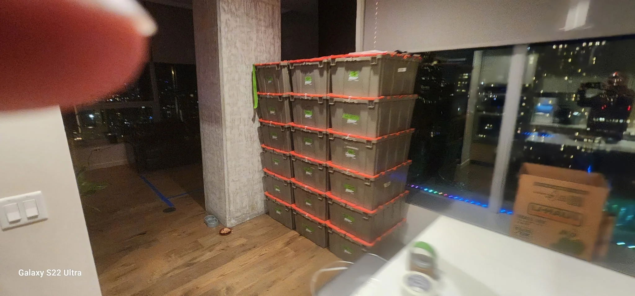 A stack of 12 plastic storage containers with orange lids arranged in a 4 by 3 grid in a room with wooden flooring and large windows at night.