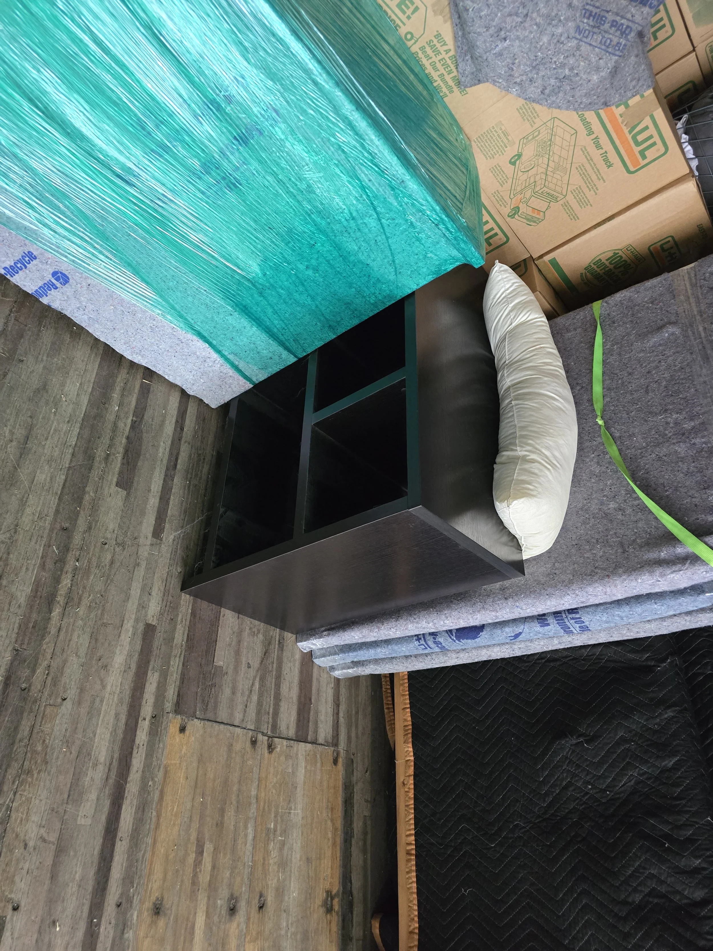 A black wooden table with empty shelf compartments, a white elongated pillow resting on top, and a silver foam board with a green tape strip on the right side. In the background, cardboard boxes and grey carpeted surfaces are visible.