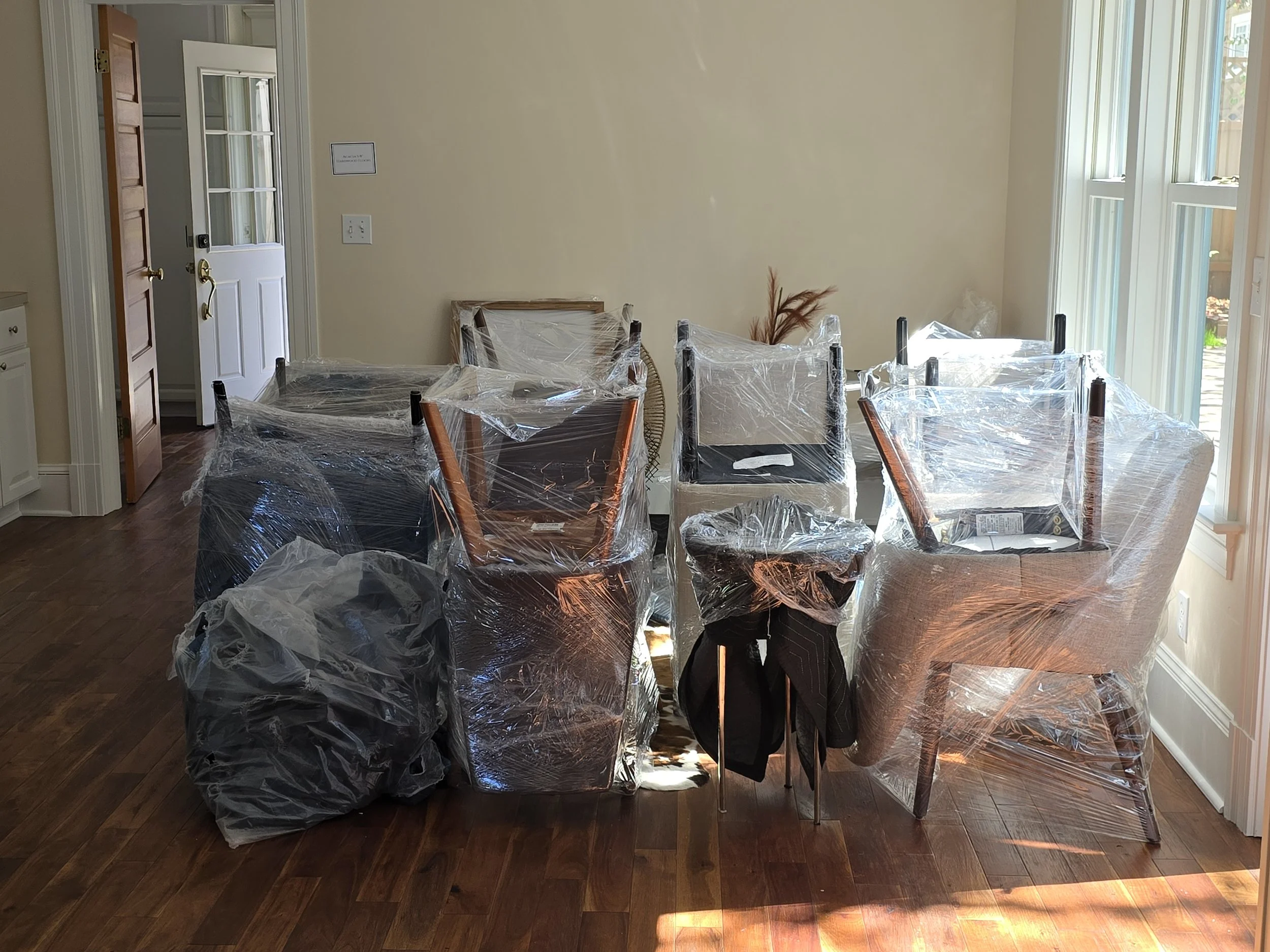 Dining room with six chairs wrapped in plastic for moving or protection, sunlight coming through windows, hardwood floor, open door to outside.