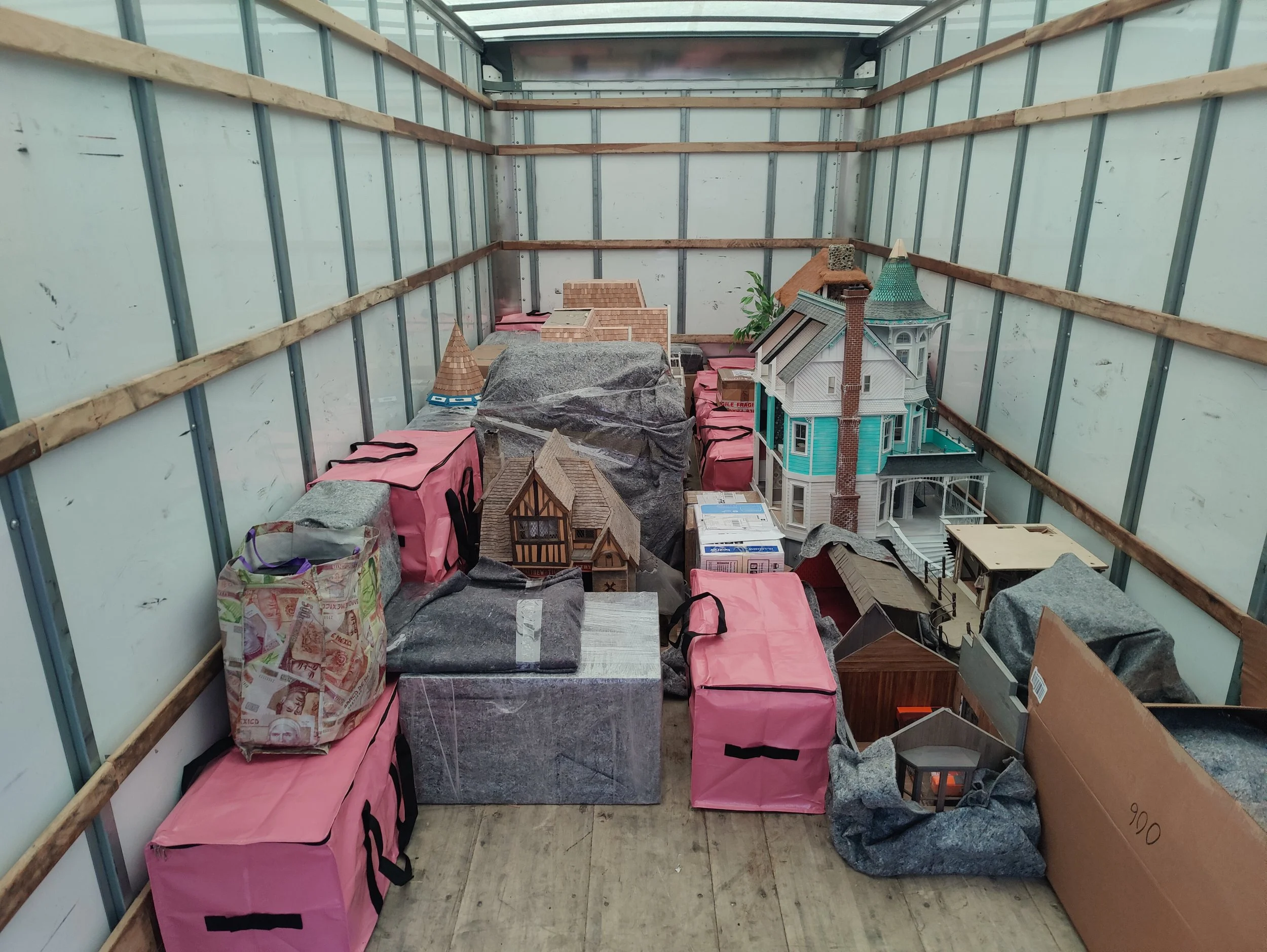 Storage container filled with dollhouses and miniature structures, some covered with protective sheets, inside a partially enclosed structure with wooden and metal framing.