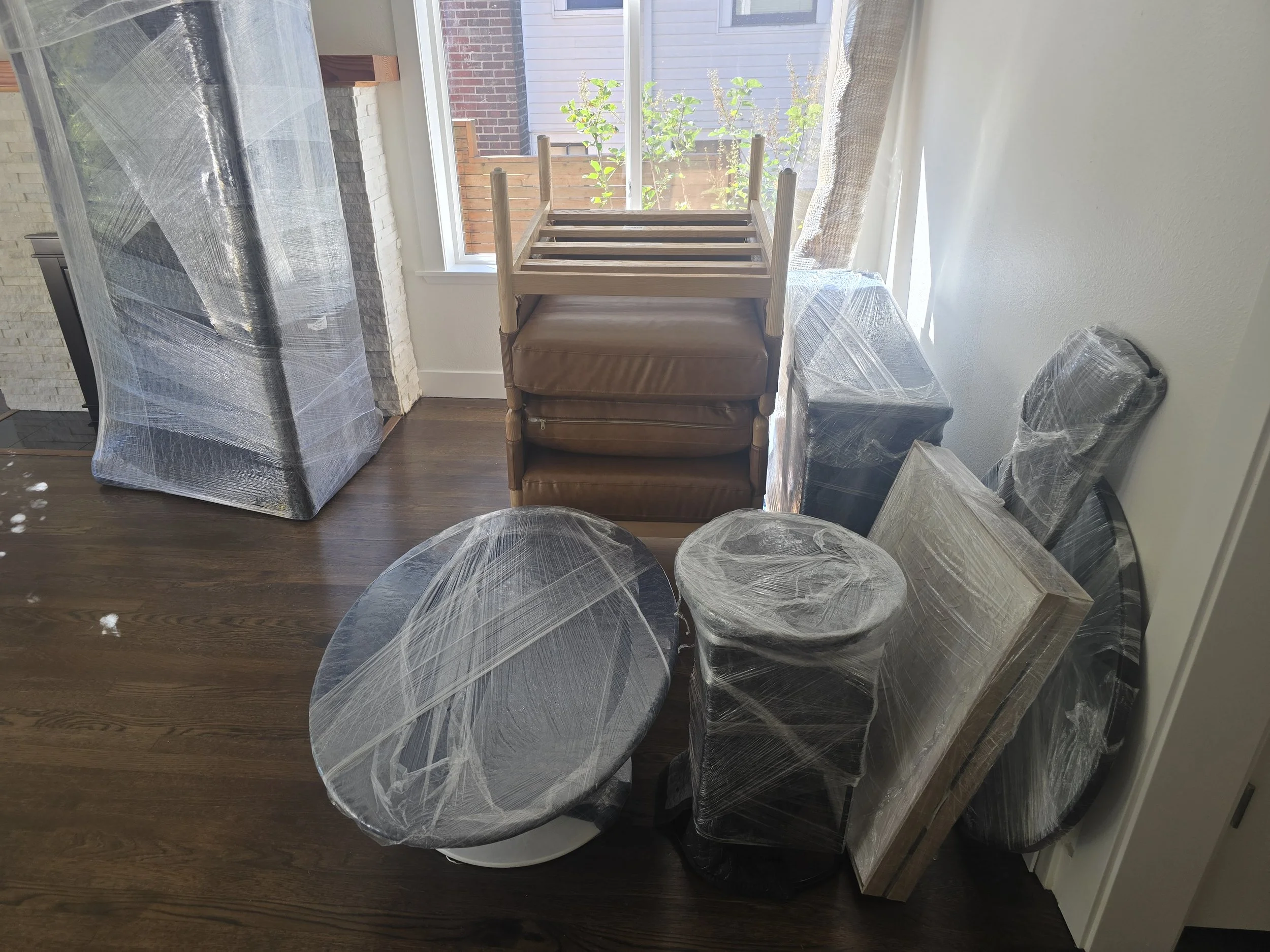 A room with furniture wrapped in protective plastic, including a round table, chairs, a dresser, a side table, and a small cabinet, with a window letting in natural light.