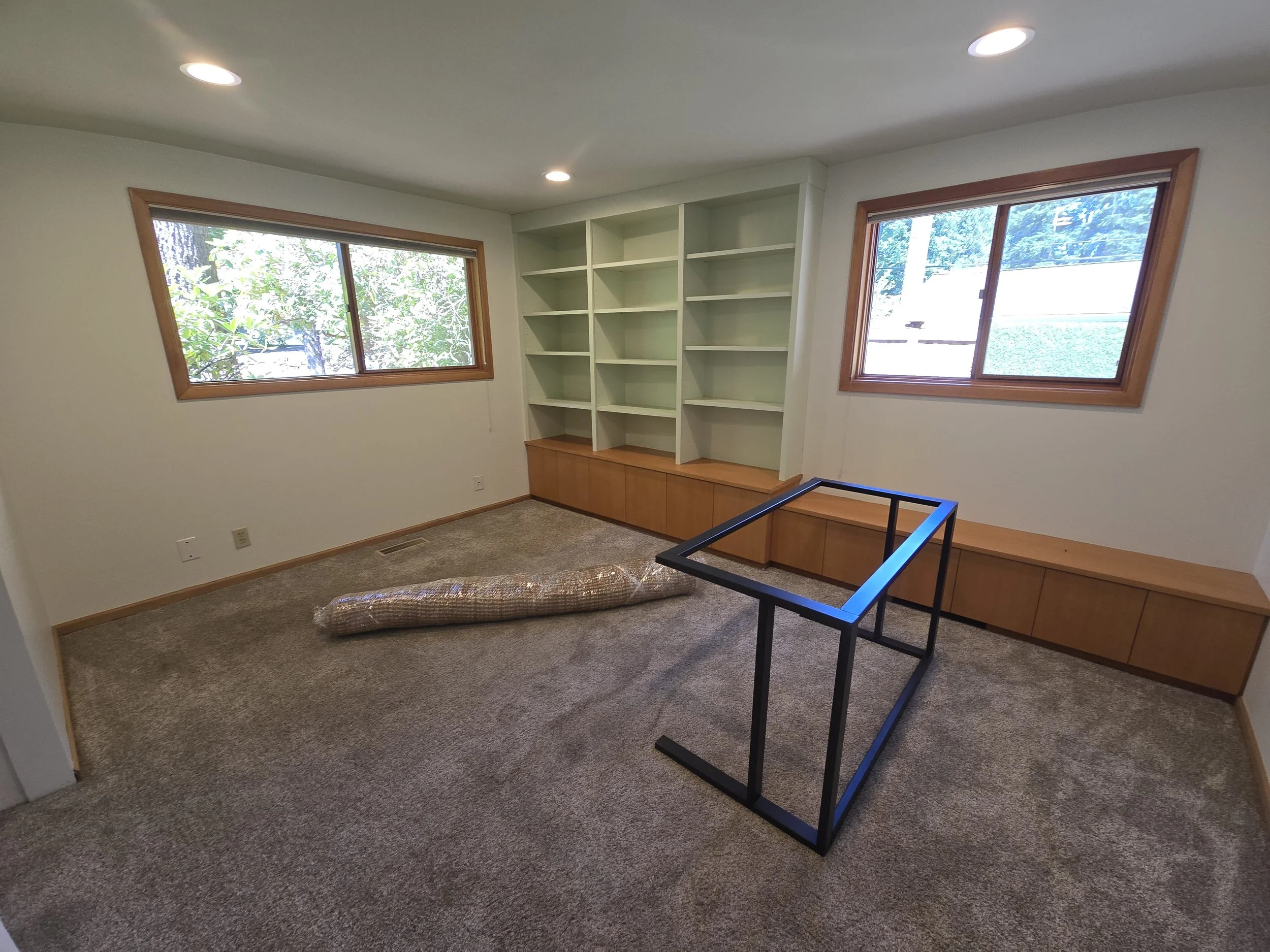 An empty room with beige carpet, two large windows, built-in white shelves with a wooden base, a black metal frame in the center, and a rolled-up carpet on the floor.