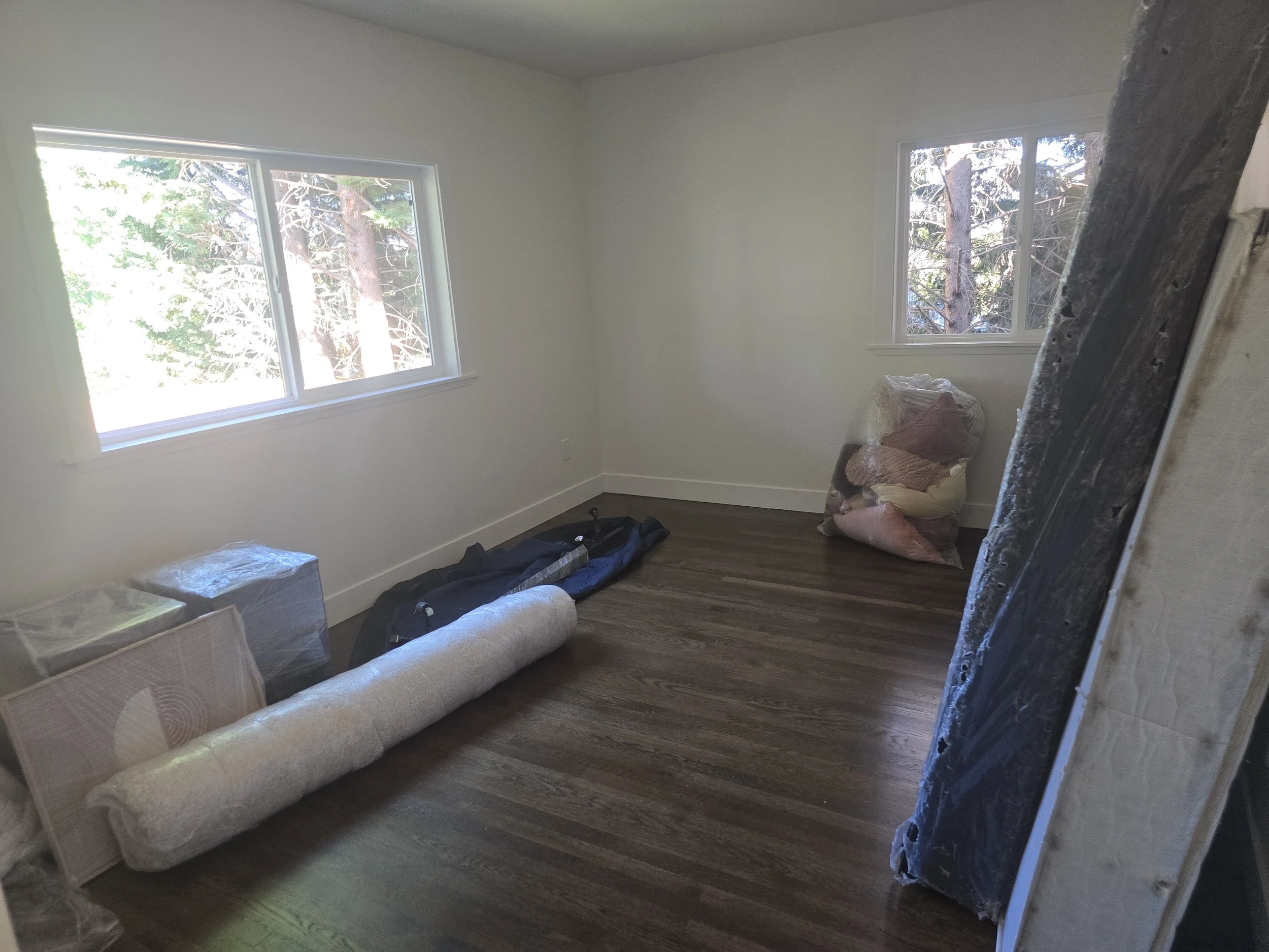 Empty room with two windows, wooden floor, and miscellaneous items including a rolled-up carpet, bags, and wrapped items, suggesting it is under renovation or moving.