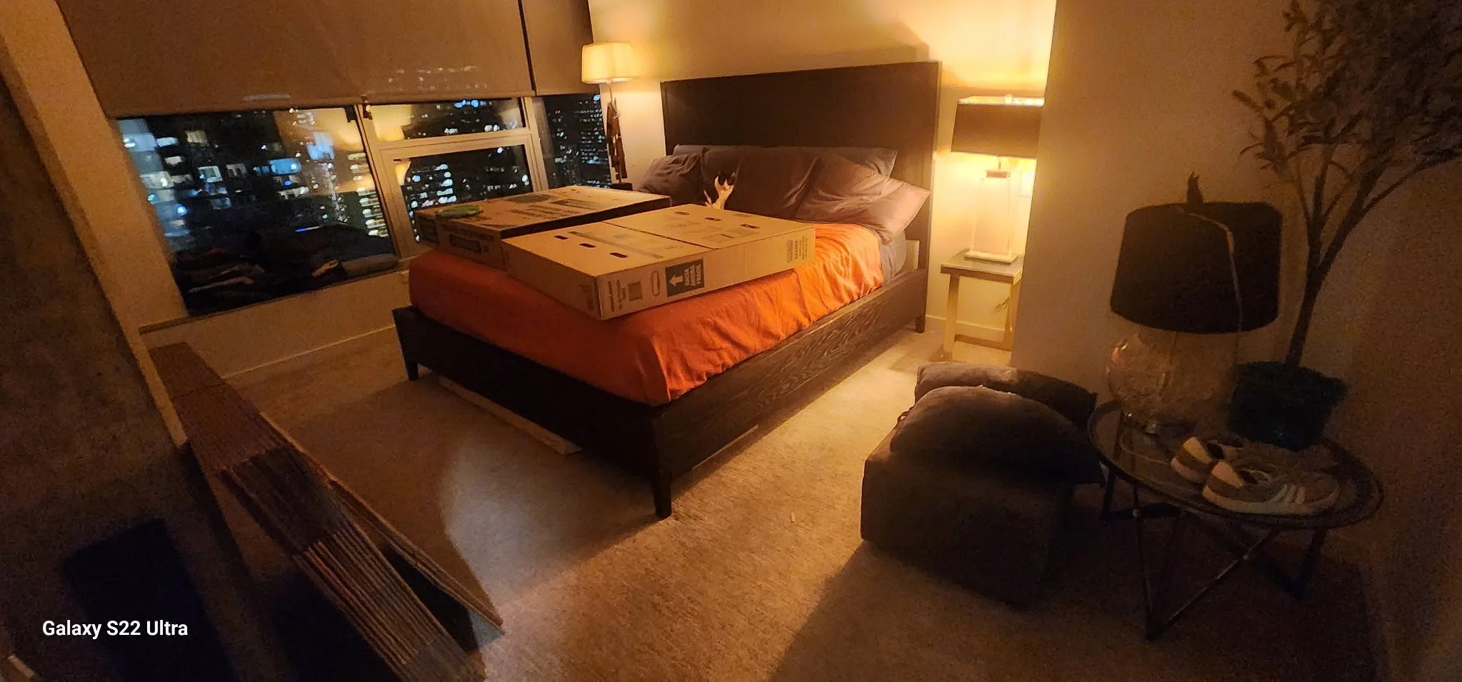 A cozy bedroom with a large bed, an orange bedspread, and a black headboard. There are boxes on the bed, pillows, a nightstand with a lamp, and a window showing city lights at night. Additional furniture includes an armchair, a side table with a lamp
