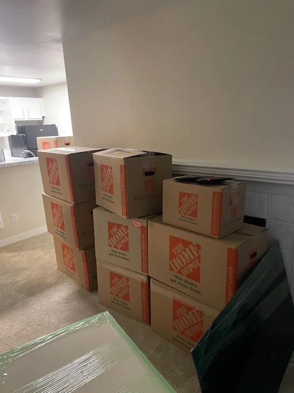 Multiple moving boxes stacked in a room next to a wall, with a kitchen area visible in the background.