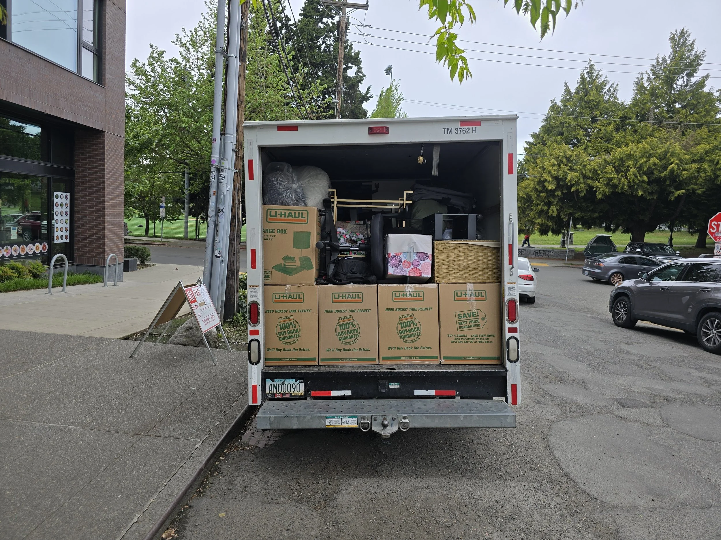 Back of a U-Haul truck filled with boxes and household items, parked on a street near a sidewalk and trees.