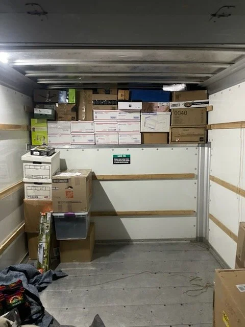 Empty delivery truck with boxes and packages stacked at the front, some shelves and items at the back, and clothing or fabric on the floor.
