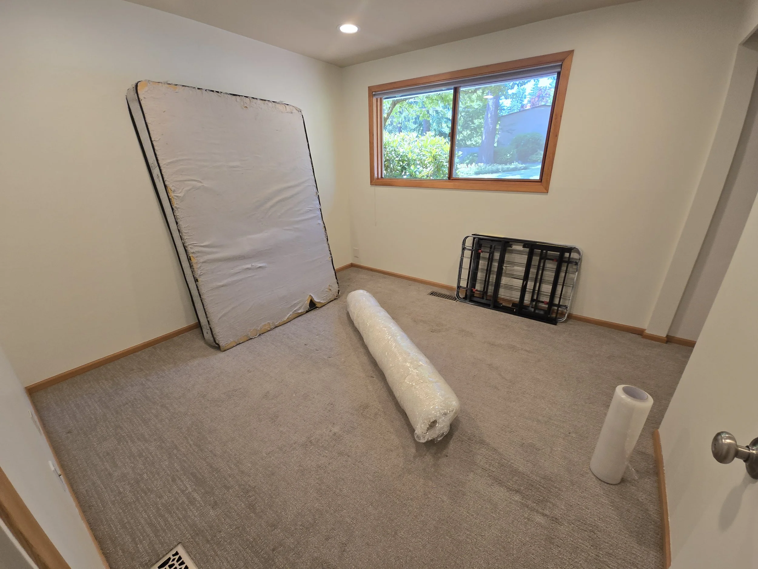 Empty room with a mattress leaning against the wall, a rolled-up plastic or foam pad on the carpet floor, a small window showing trees outside, and a folded bed frame leaning against the wall.