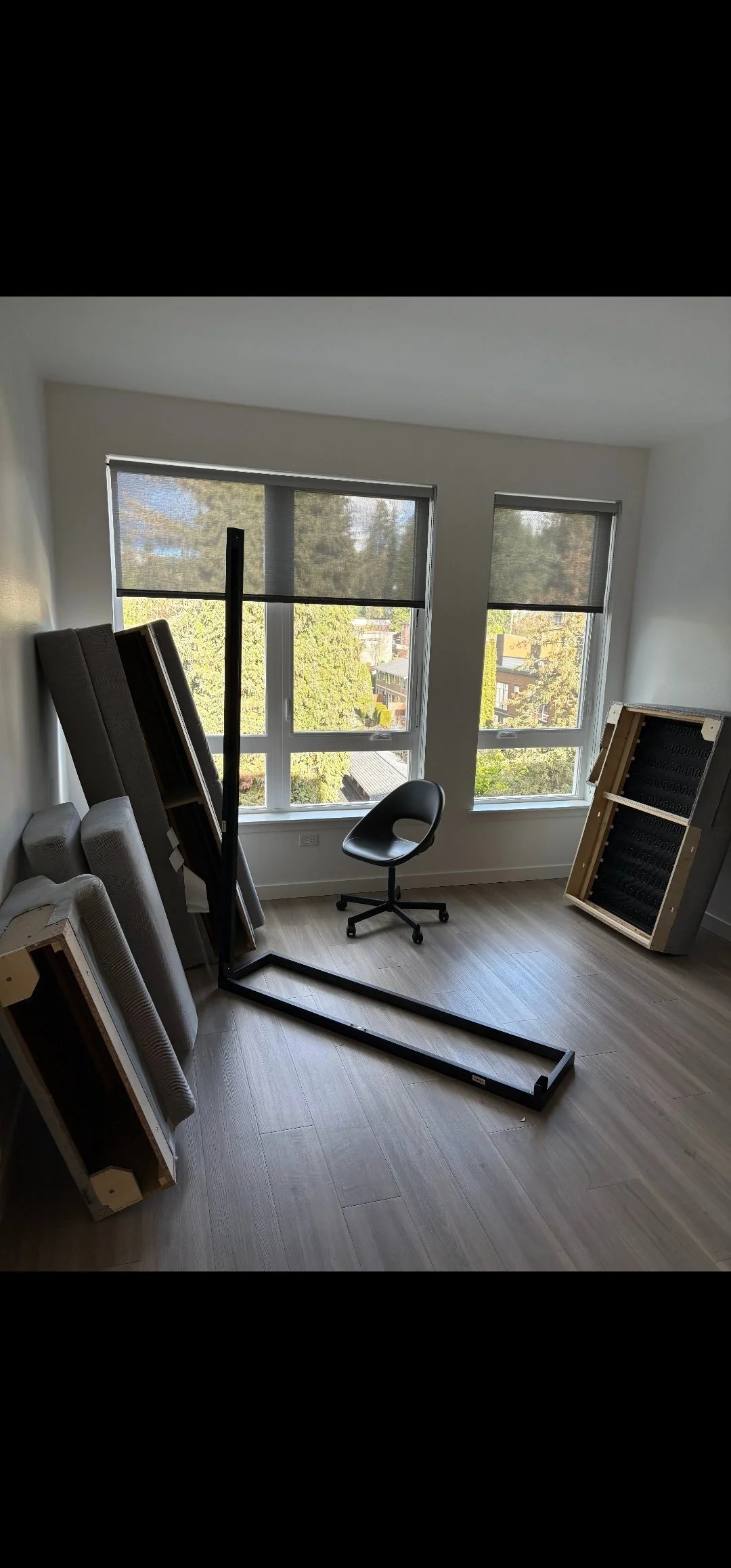 Unfurnished room with three large windows, a black office chair, a dismantled bed or shelving, and stacked boxed furniture, with trees outside the windows.