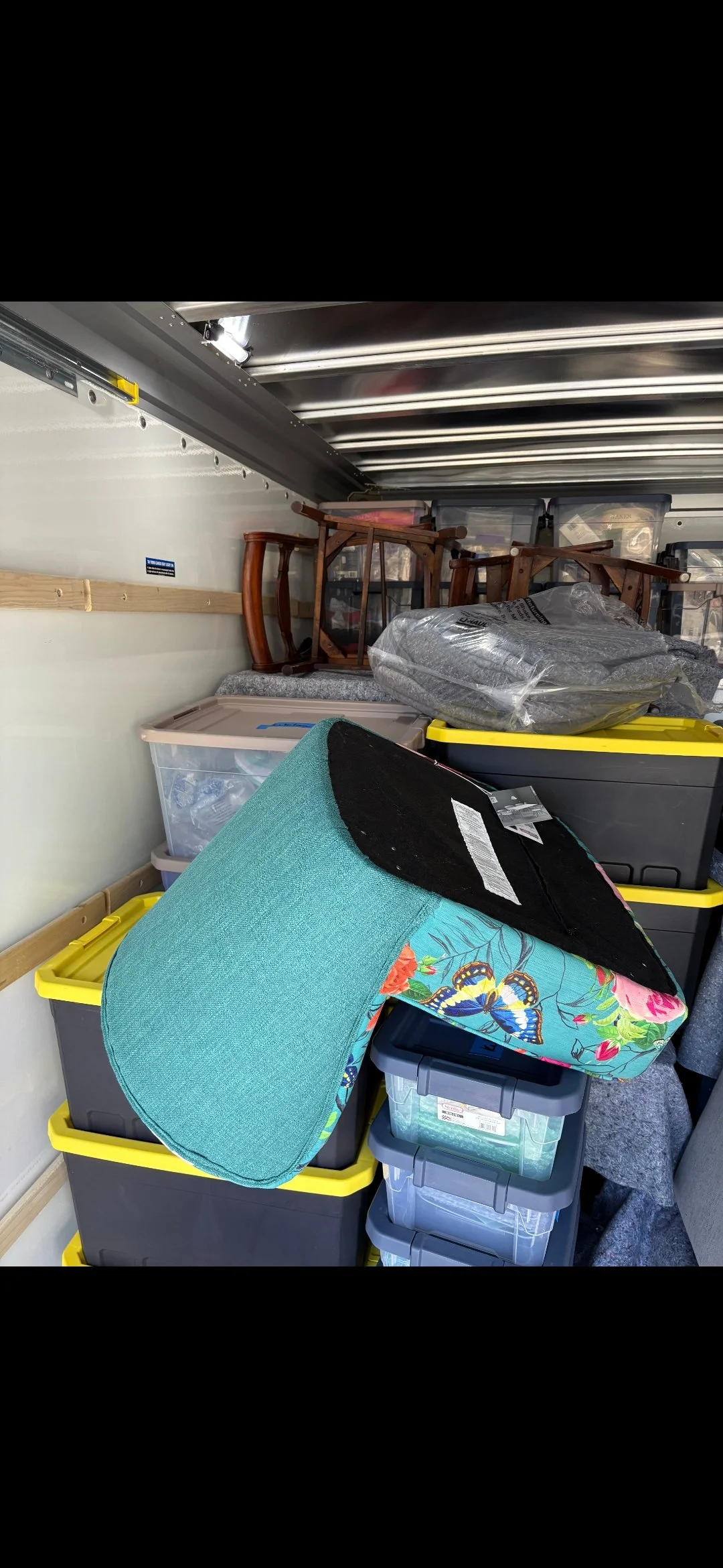 Moving boxes and furniture packed in a truck, including plastic storage bins, a floral armchair cushion, and wooden chairs.
