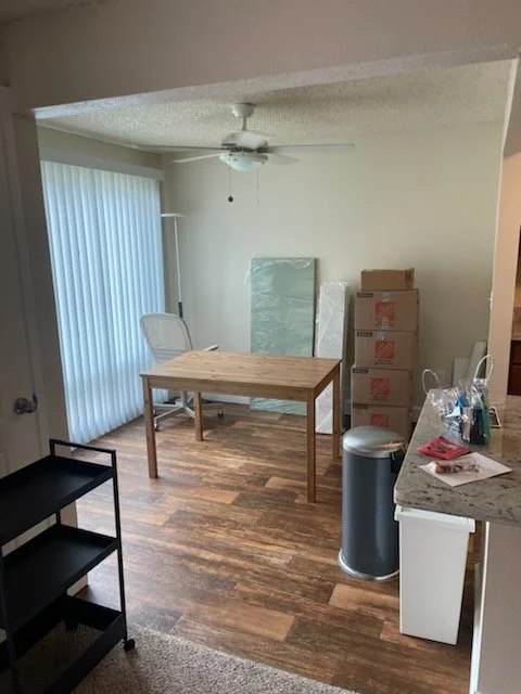 A partially furnished dining or workspace area with a wooden table, a white chair, and several stacked boxes in the corner. There's a ceiling fan, a window with vertical blinds, and various items on the countertop, including a bottle and paper.