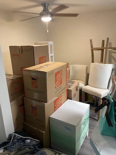 Room with stacked moving boxes and white chairs, ceiling fan, and a small table with bags.