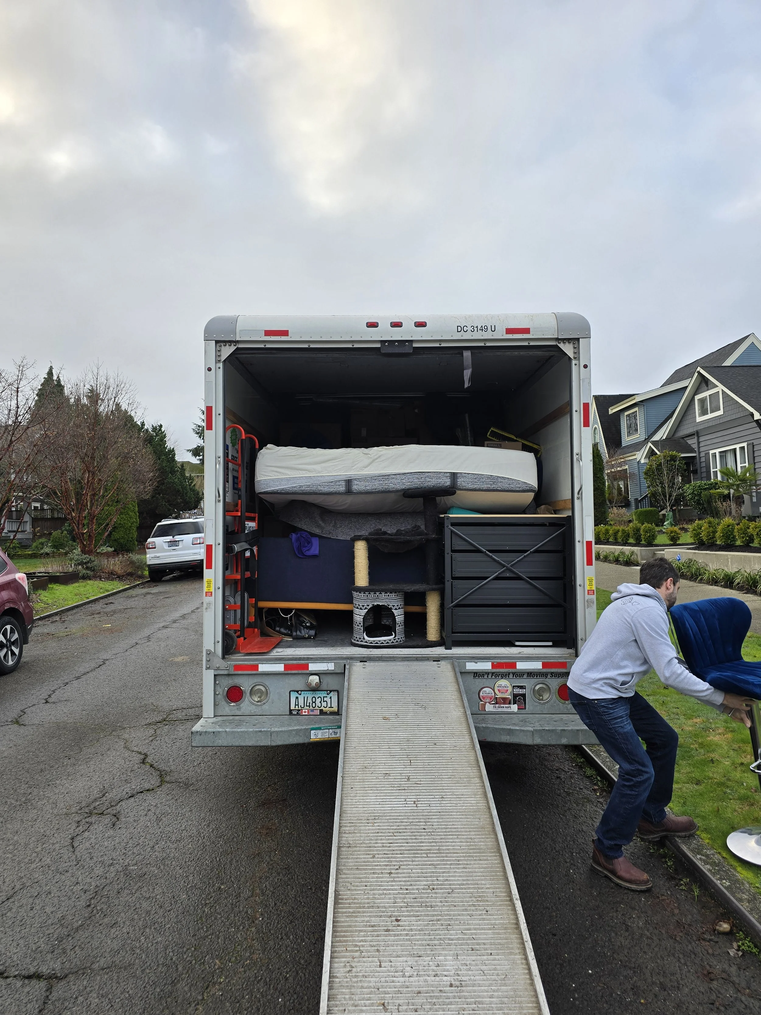 Person loading a moving truck with furniture and household items on a residential street.