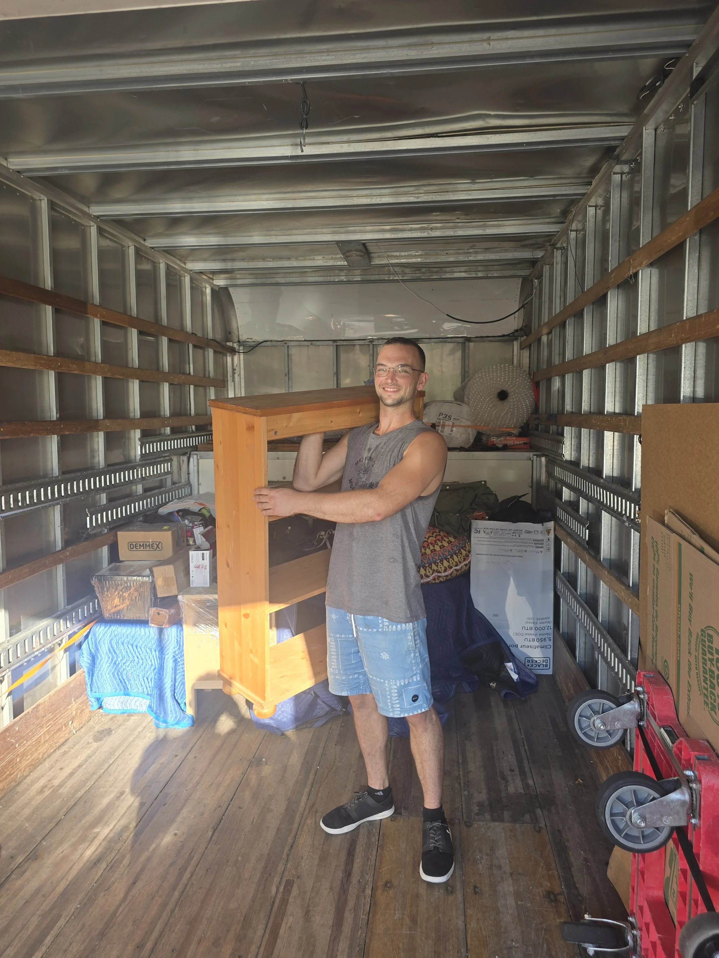 A man lifting a small wooden bookshelf inside a storage unit, smiling, wearing a sleeveless shirt and shorts.