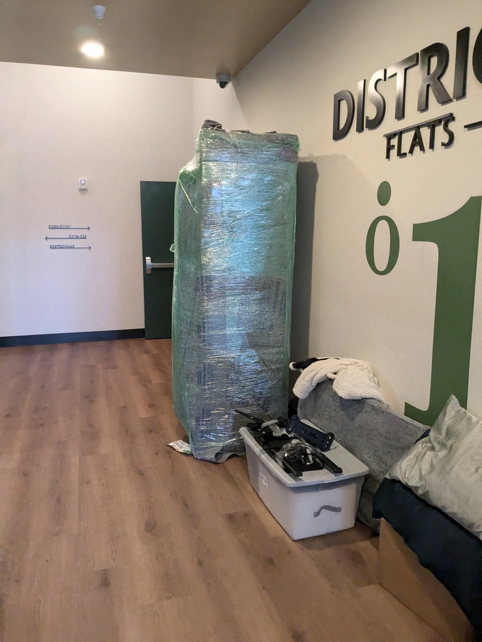 Wrapped furniture or large item in plastic, placed next to a bench with a pillow and blanket, in a hallway with sign indicating district flats and directions to restrooms.
