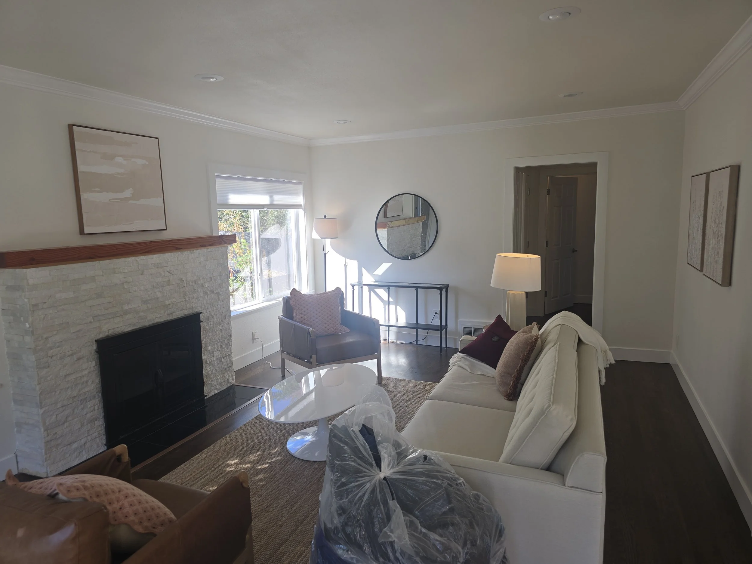 Living room with fireplace, white sofa, leather armchair, decorative pillows, windows, floor lamps, wall art, round mirror, black console table, and light-colored walls.