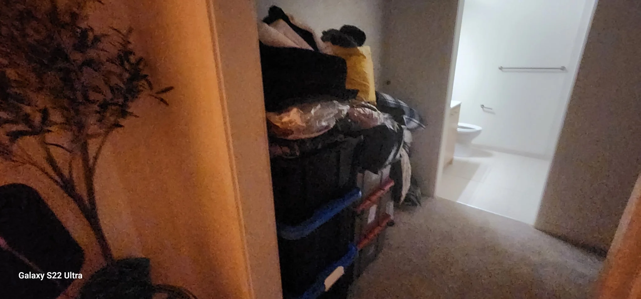 A cluttered room with a wooden table, a pile of bags and clothes, and a view of a bathroom with a toilet in the background.
