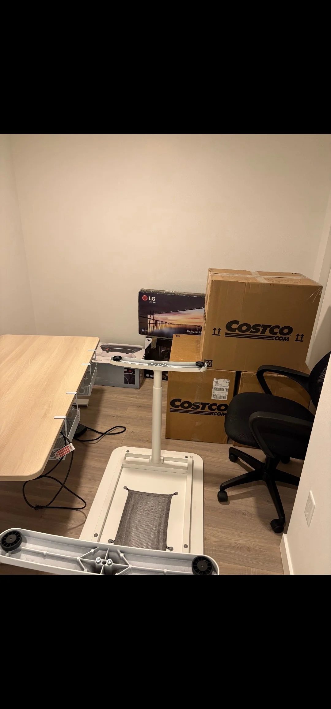 Room with boxed and unboxed electronics and furniture, including a large cardboard box labeled Costco, a flat-screen TV, a chair, and part of a disassembled standing desk or equipment.