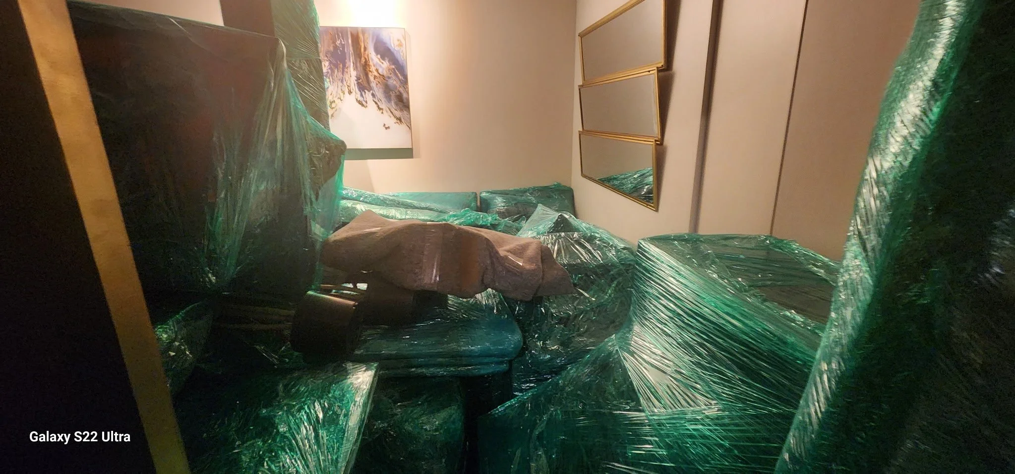 Living room with furniture wrapped in green plastic, including sofa and a chair. There is a piece of furniture with a cloth on it and a wall with decorative mirrors and framed art.