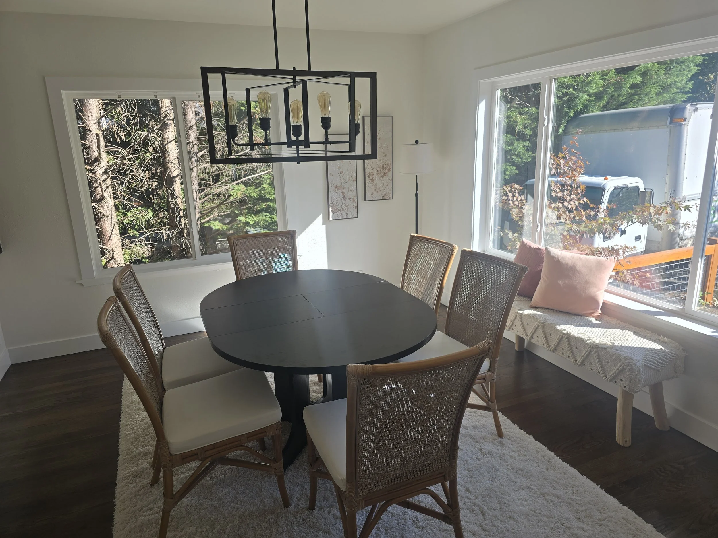 Dining room with a round black table, six wicker chairs, large windows showing trees and a truck outside, a white bench with pillows, modern chandelier, and art on the wall