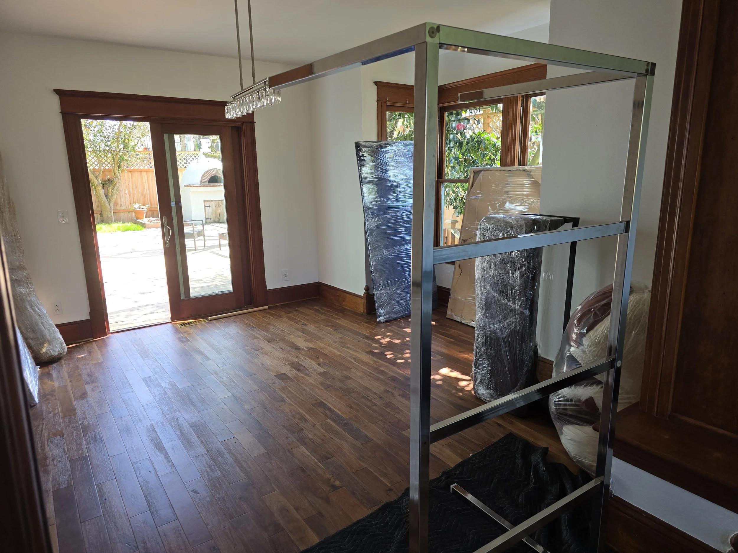 Empty room with hardwood floors, sliding glass door leading to outdoor patio, and large windows showing garden. A metal frame structure and wrapped furniture are present inside.