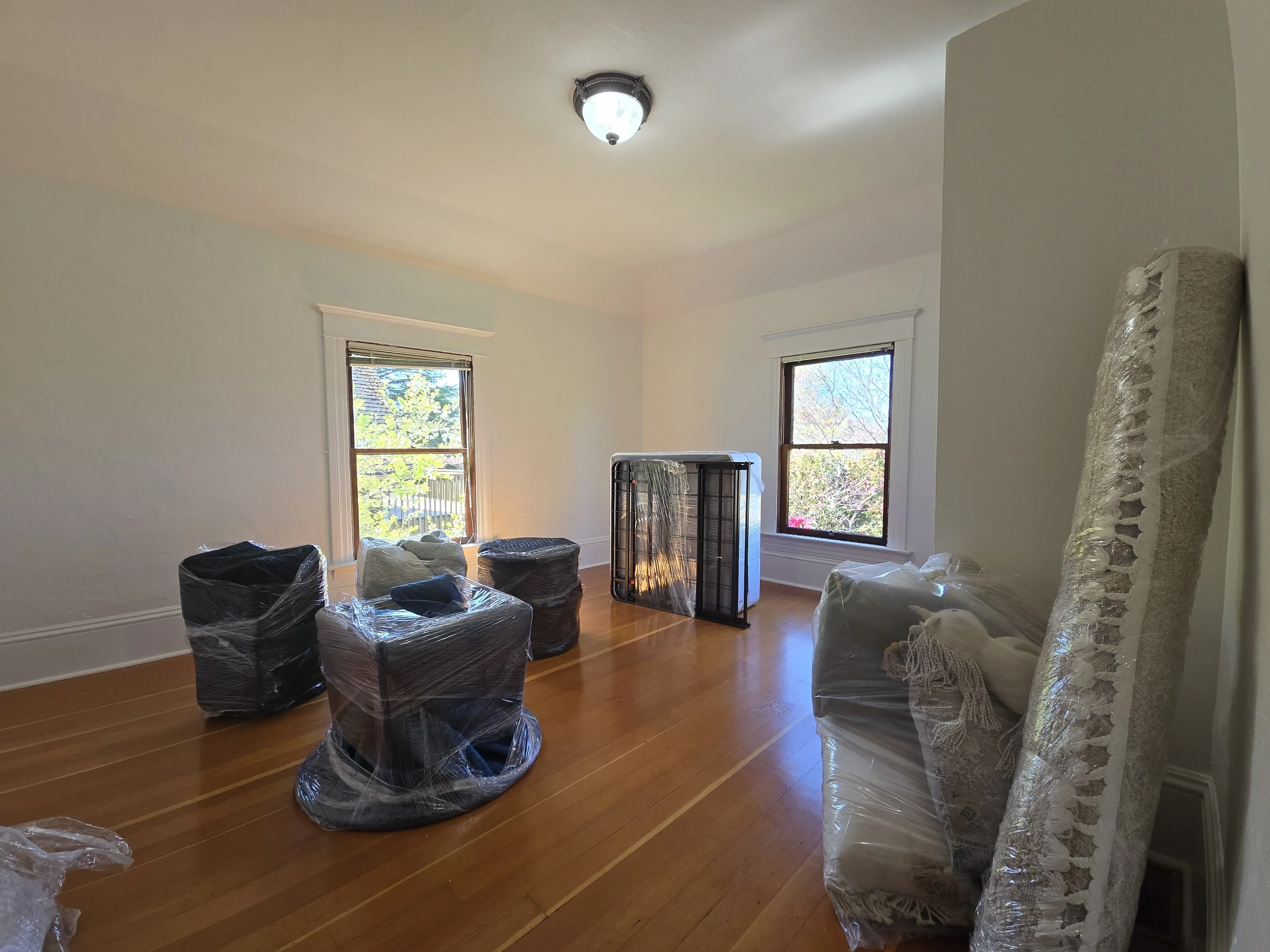 Empty room with hardwood floors, two windows, and a ceiling light fixture. Several furniture pieces wrapped in plastic are stacked or leaning in the room, likely for moving or renovation.