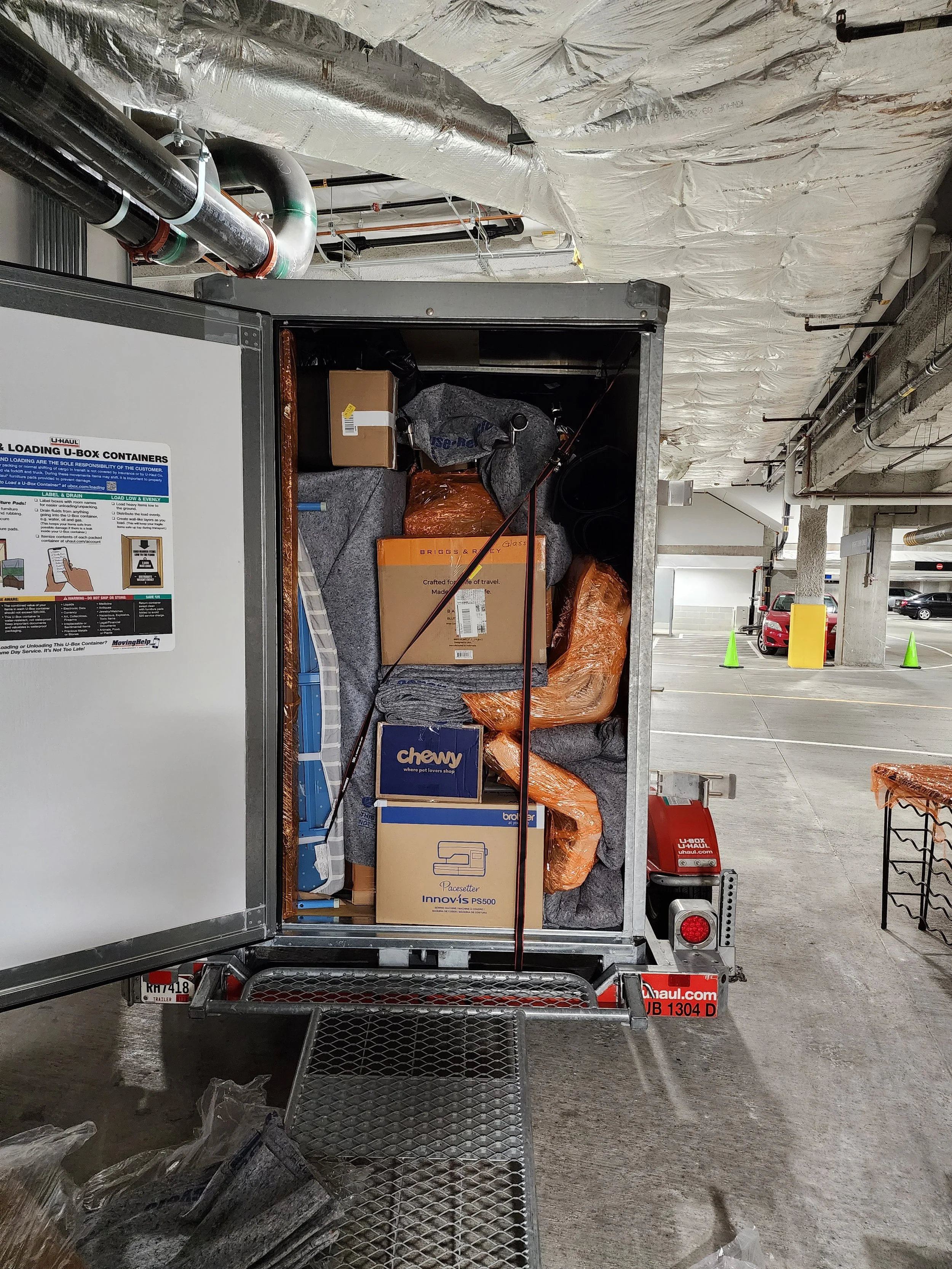 Back of a loaded U-Haul trailer filled with boxes, blankets, and miscellaneous items in a parking garage, with cars and orange cones in the background.
