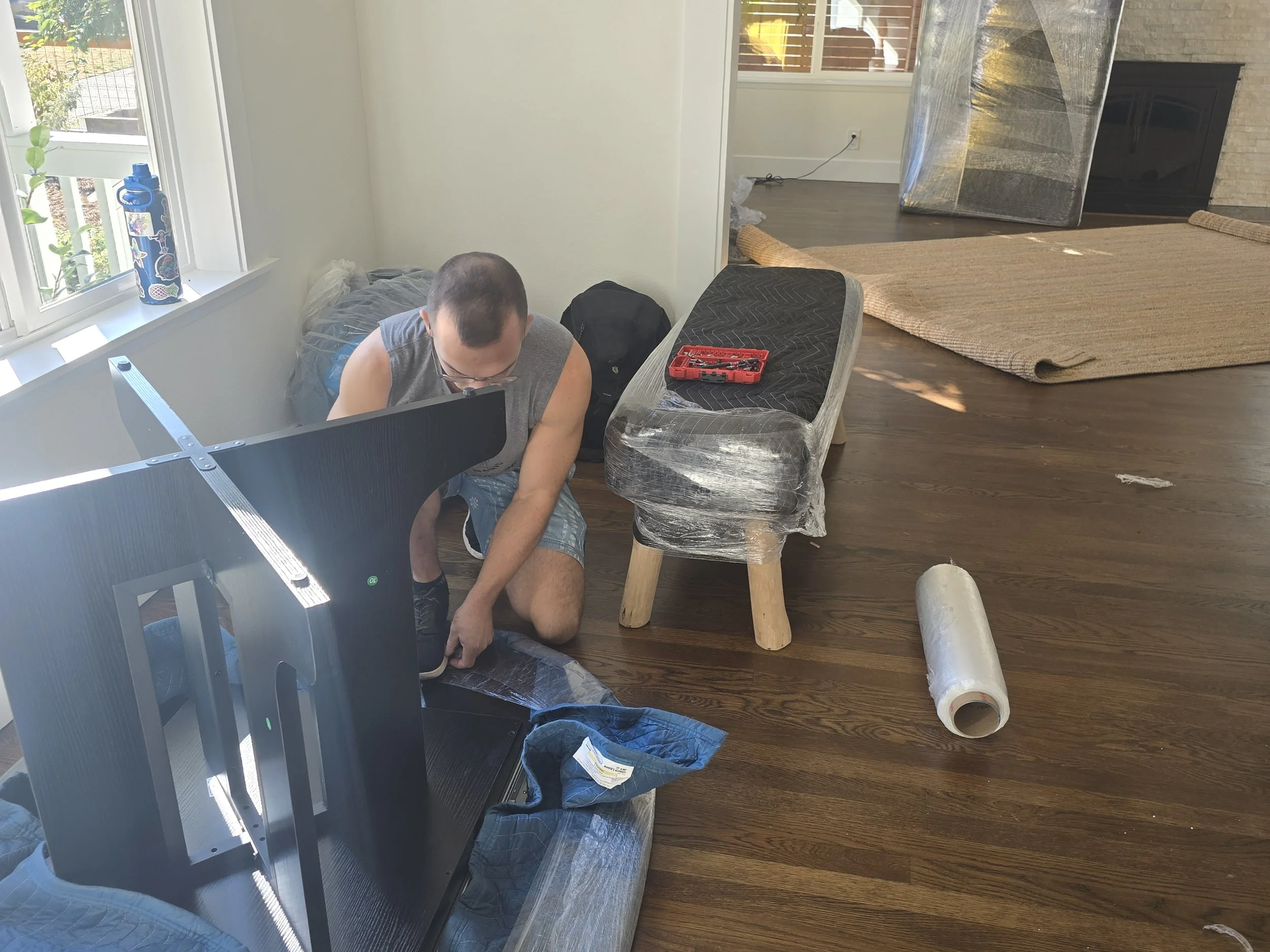 A man installing or repairing a large flat-screen TV or monitor in a living room. He is kneeling on the floor next to the TV, which is lying face down. There are tools on a small stool nearby, and a roll of plastic wrap or protective film on the floo