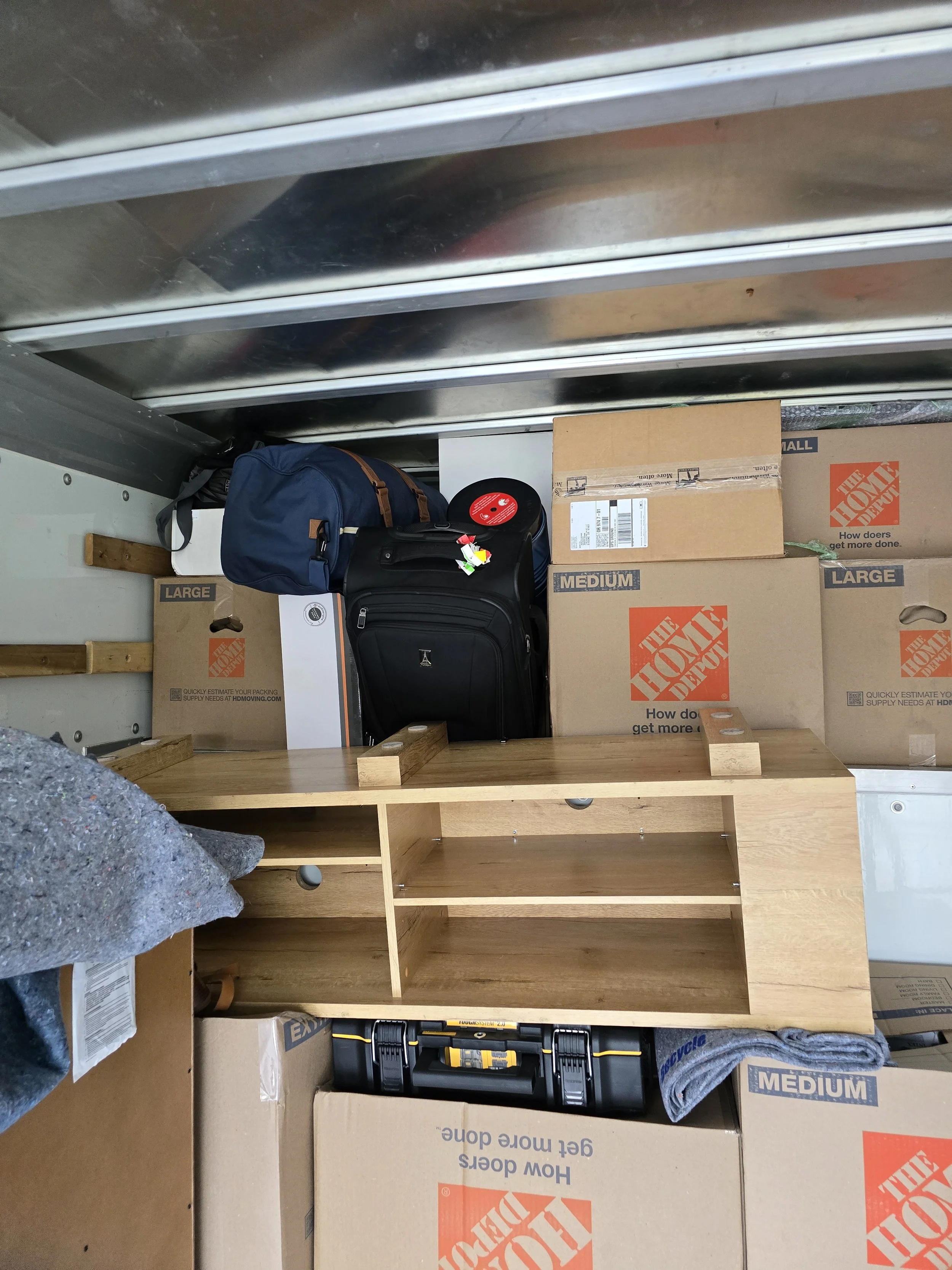 The interior of a moving truck with packed boxes, backpacks, a wooden shelf, and a toolbox.
